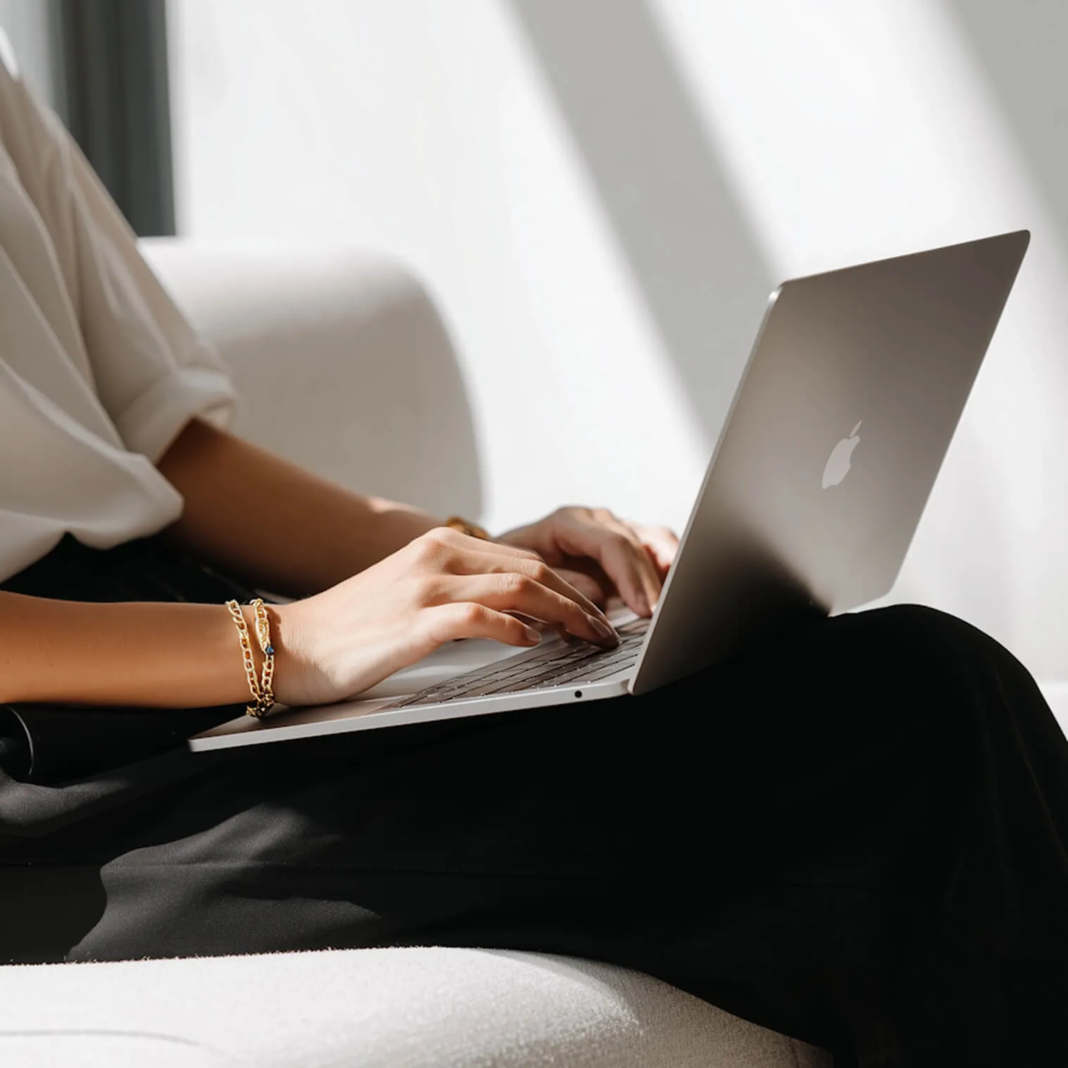 Person wearing a gold bracelet typing on a MacBook laptop while sitting on a light-colored couch.
