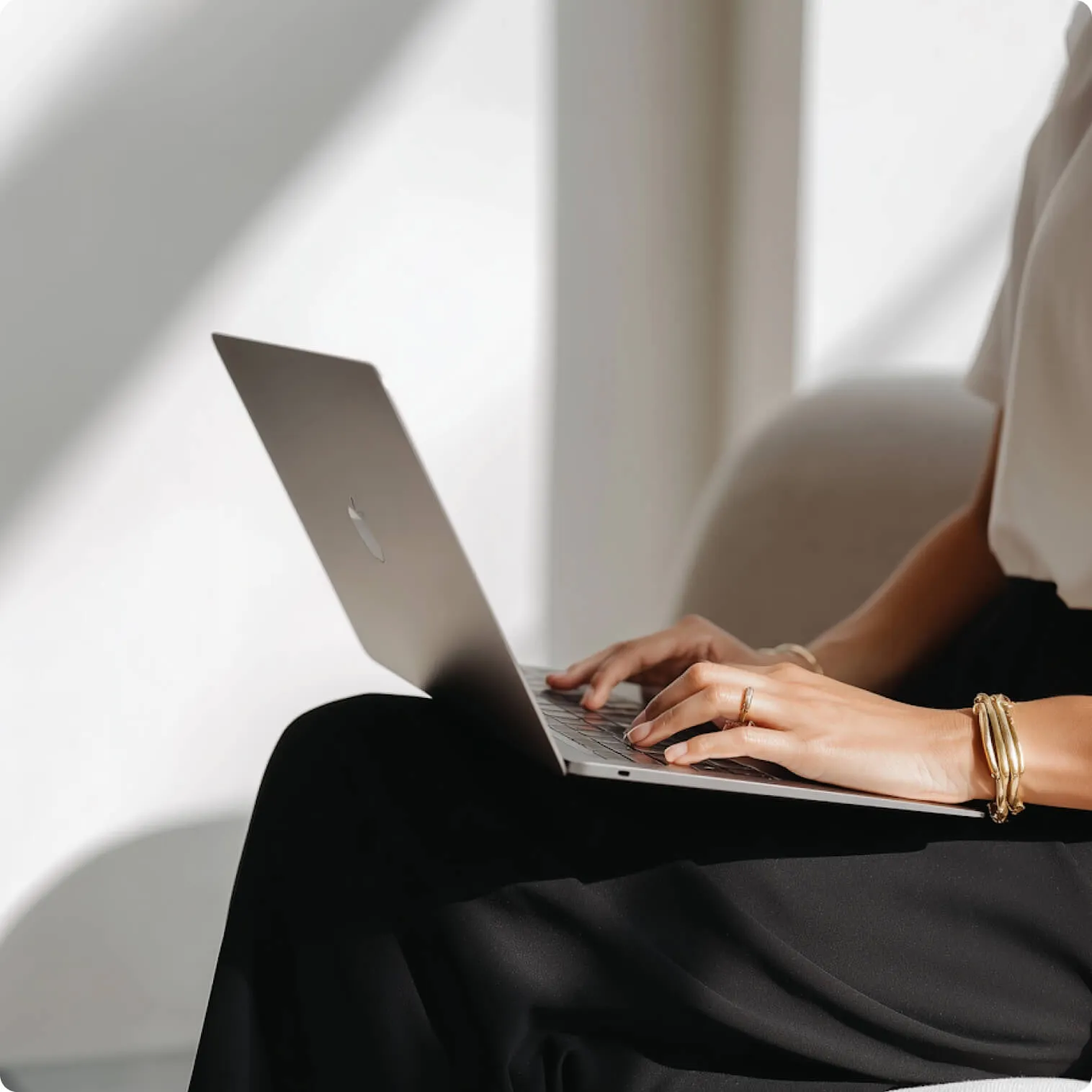 Person typing on a silver laptop while sitting, wearing a white top and black pants with gold bracelets on wrist.