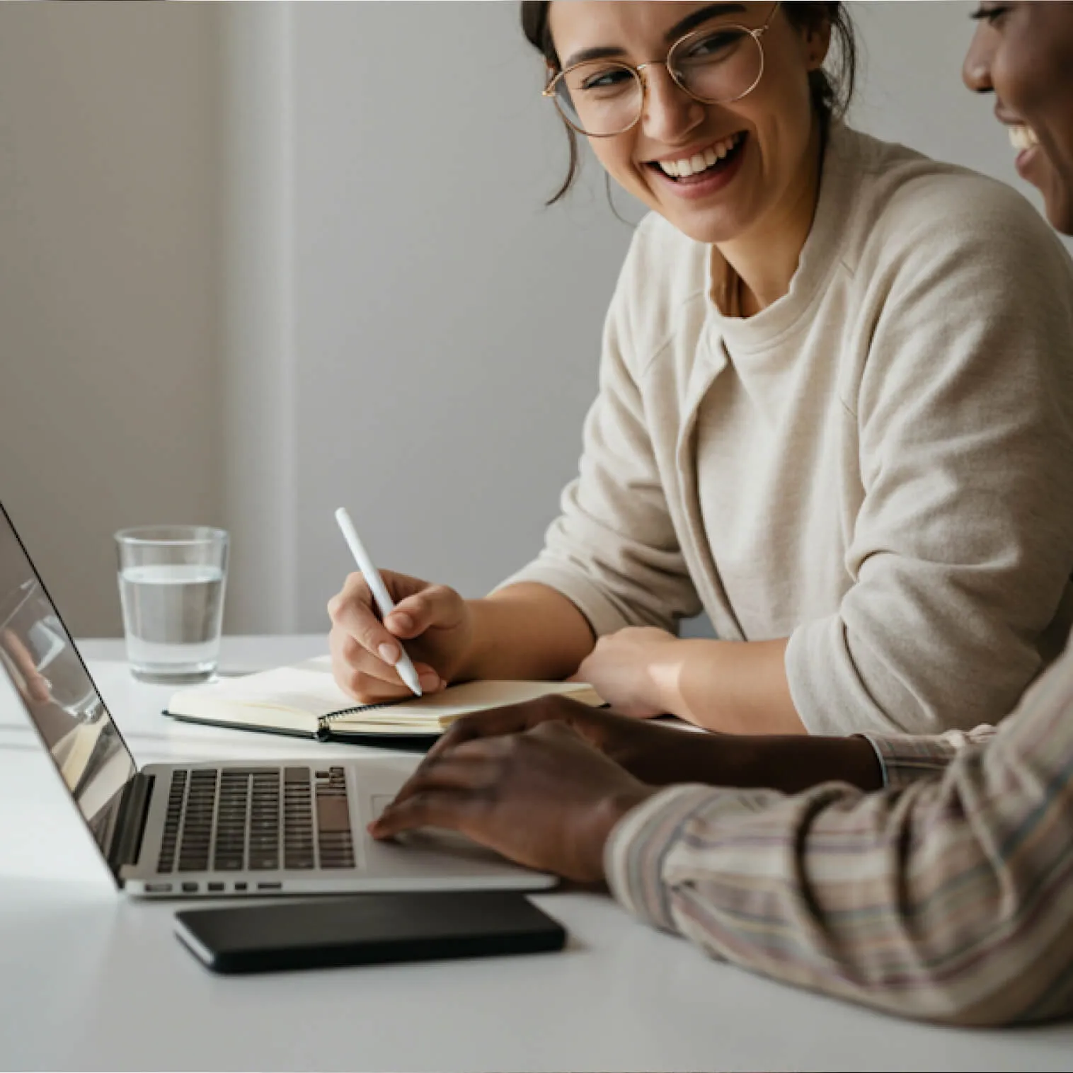 Two people collaborating at a desk with a laptop, notebook, and glass of water, smiling and writing notes.
