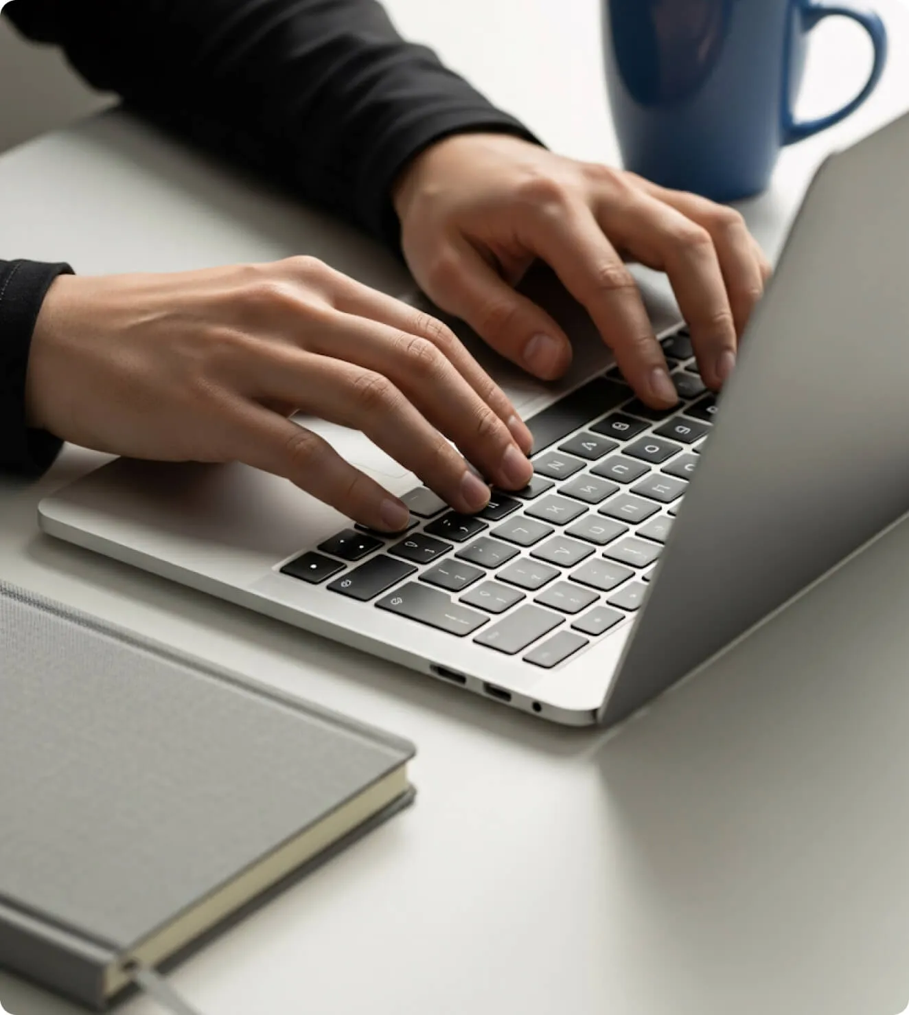 Person typing on a laptop keyboard with a blue mug and a closed notebook on the white desk.
