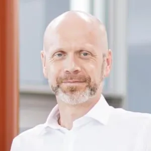 Middle-aged bald man with a light beard wearing a white shirt, smiling slightly in a bright indoor setting.