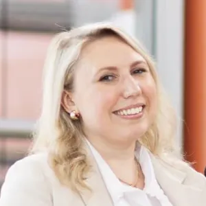 Smiling woman with blonde hair wearing a white blouse and earrings in an indoor setting.