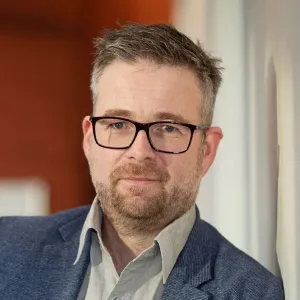 Man with short hair and beard wearing glasses and a blue blazer leaning against a wall.