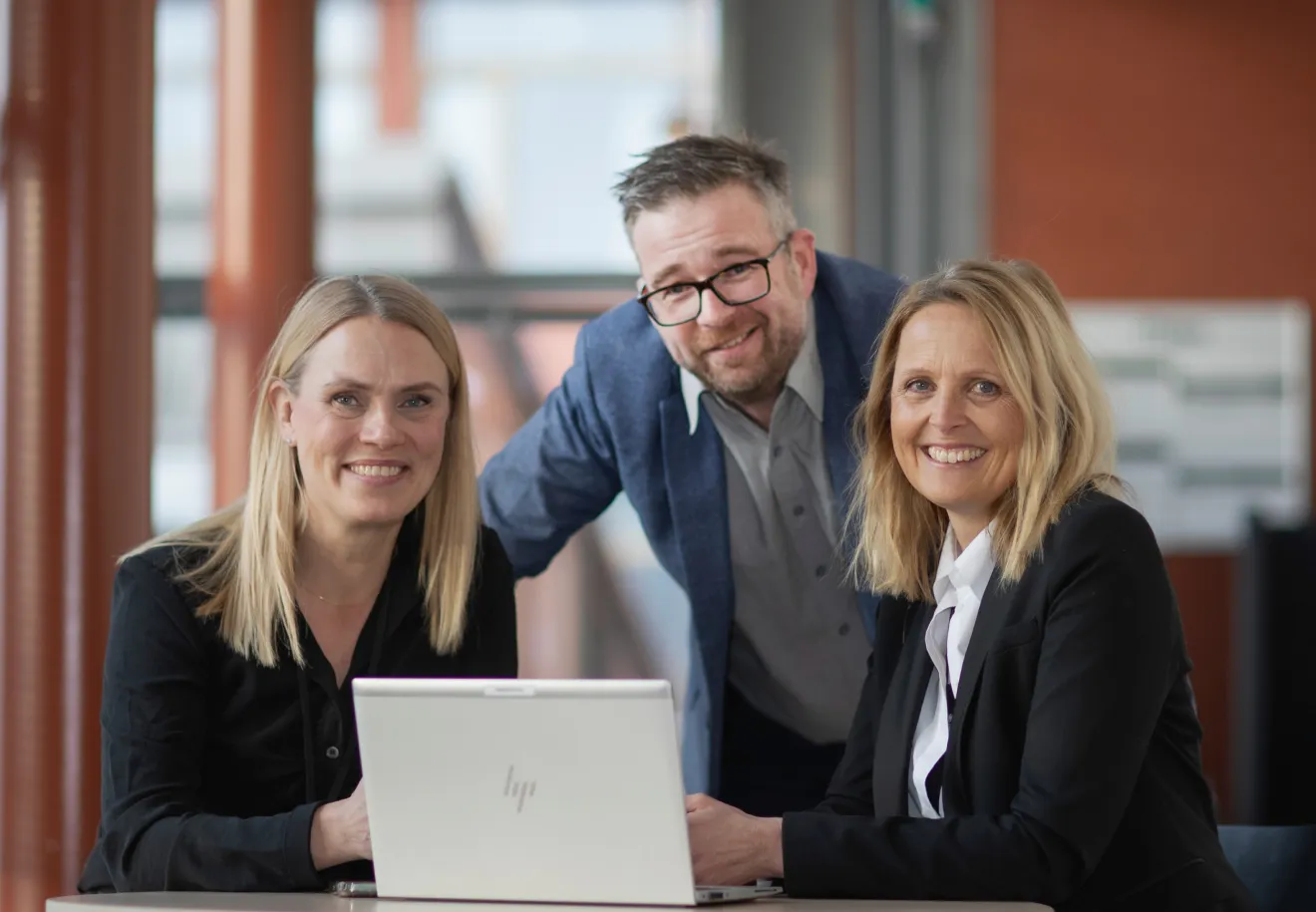 Two women and a man smiling at camera around a laptop in a modern office setting.
