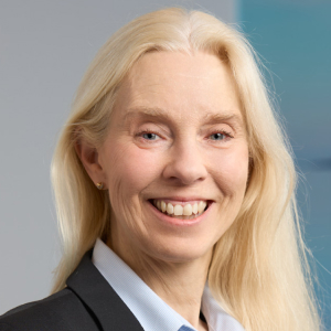 Smiling woman with long blonde hair wearing a black blazer and blue shirt against a blurred background.