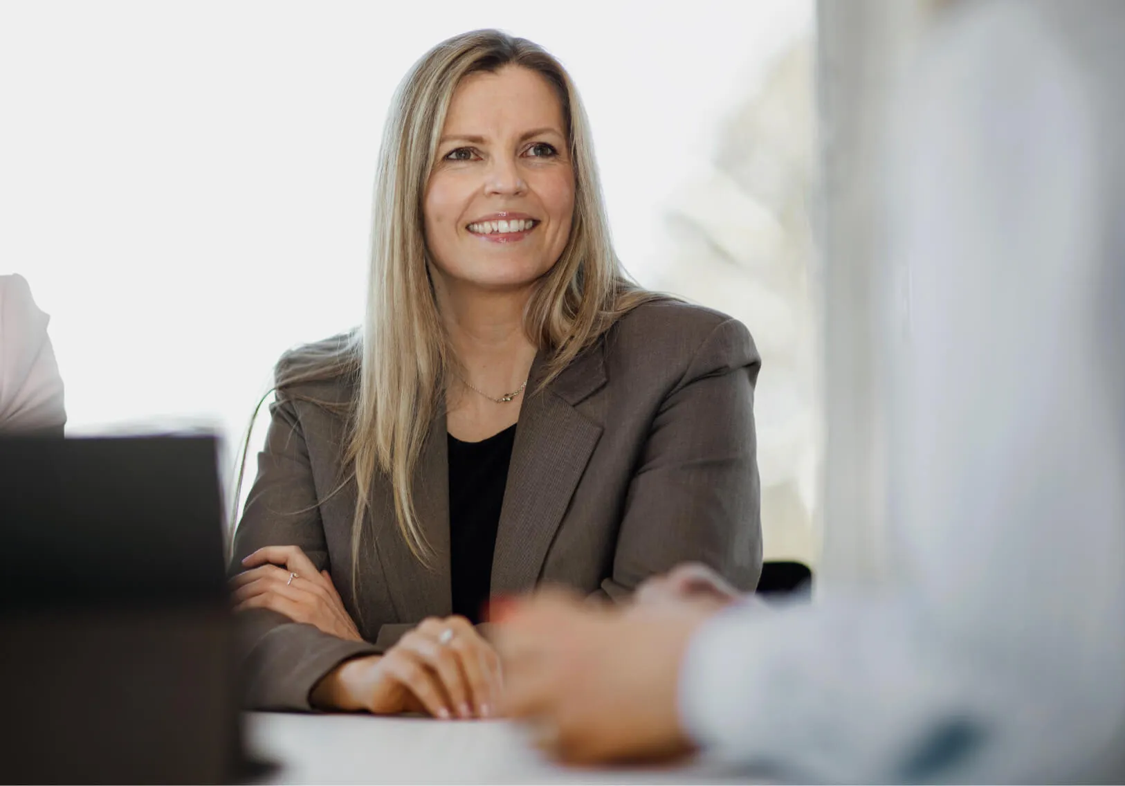 Smiling woman in a brown blazer sitting at a table in a bright meeting room.