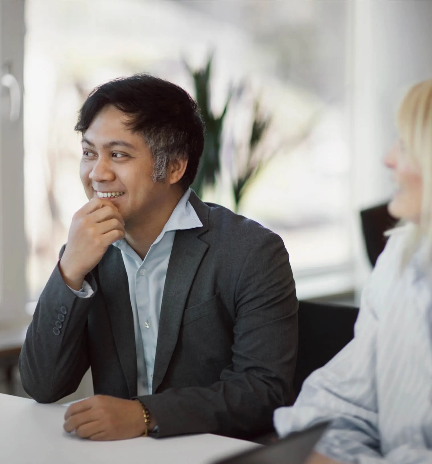 Man in gray suit smiling and sitting at a table in a bright office with a woman nearby.