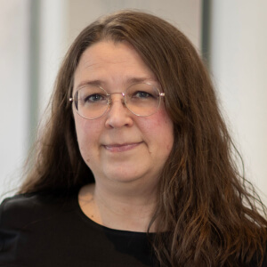 Woman with long brown hair and glasses, wearing a black top, smiling softly.