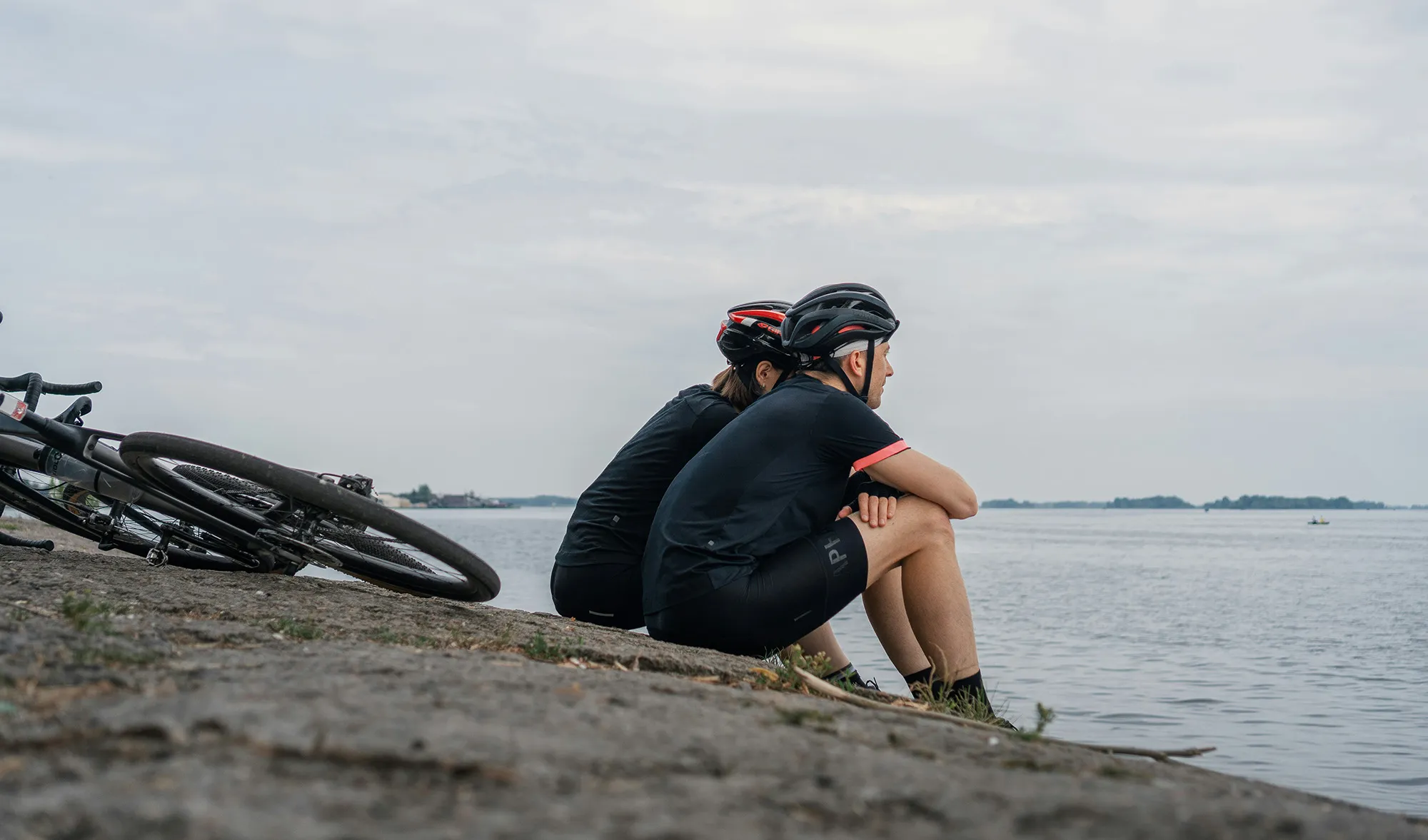 Two cyclists in helmets resting sitting on rocky shore facing calm water under cloudy sky.