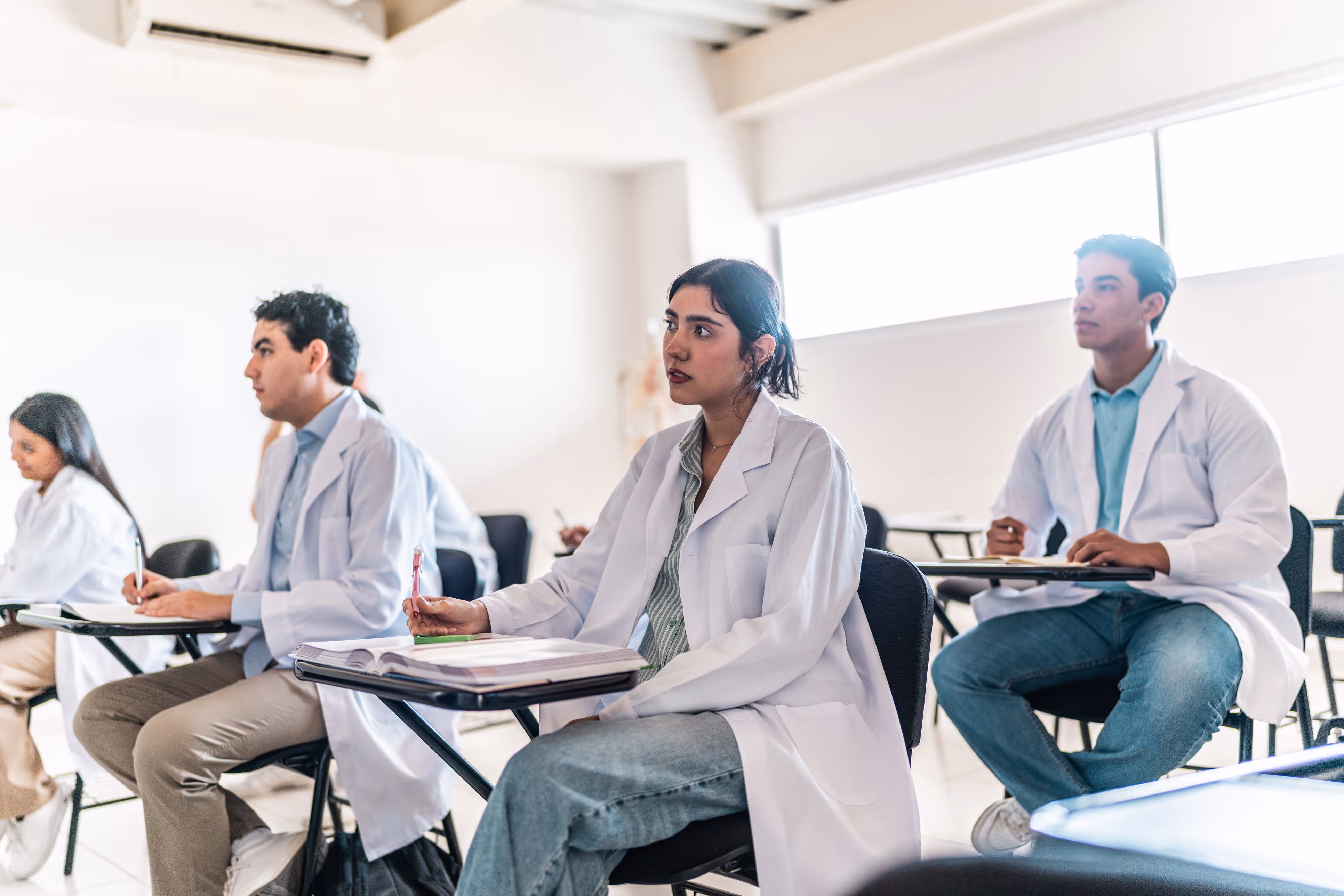 Medical students wearing white coats sit attentively in a bright classroom with notebooks and pens.