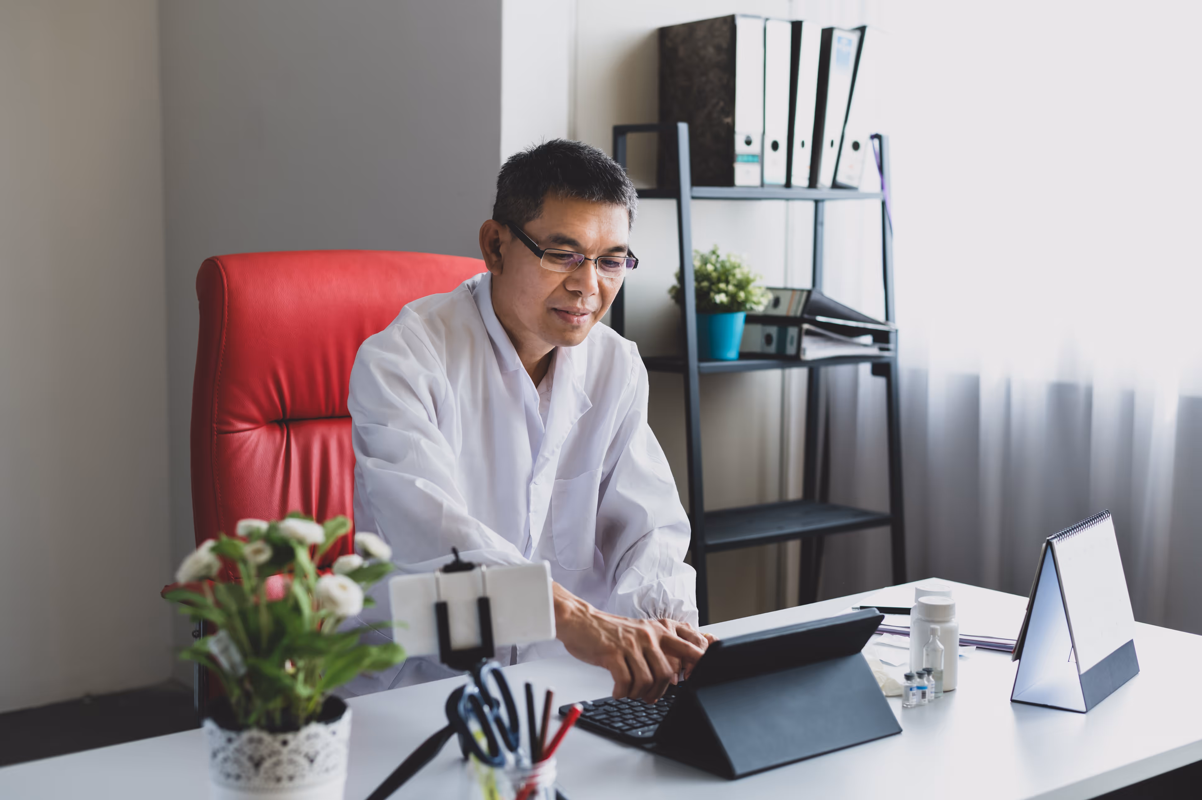 Man in white coat sitting in a red chair, using tablet at a desk with calendar and plants.
