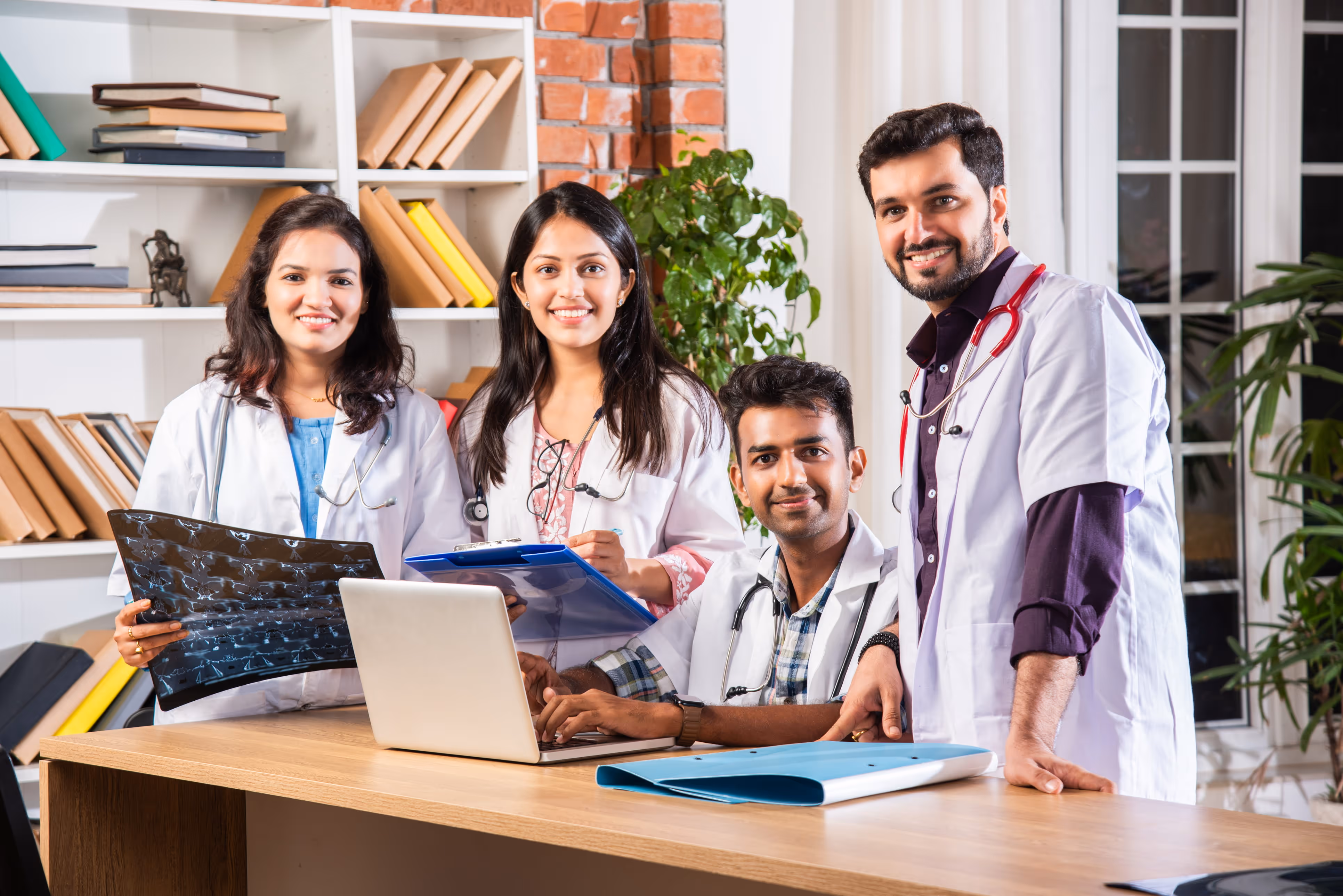 Four doctors in white coats with stethoscopes around a table with laptop and medical files.