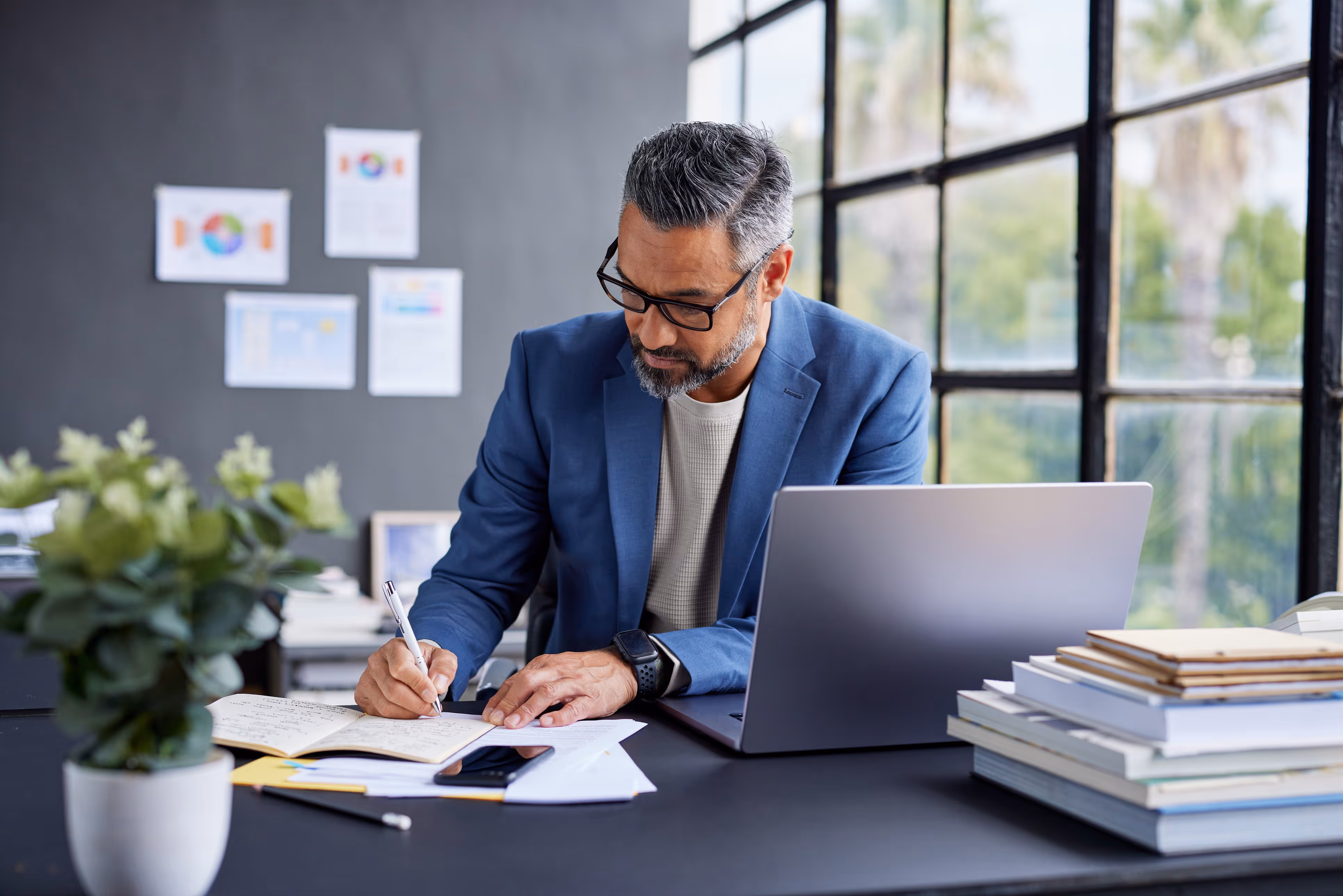 Man with glasses writes notes at desk with laptop, books, and plant near large window.