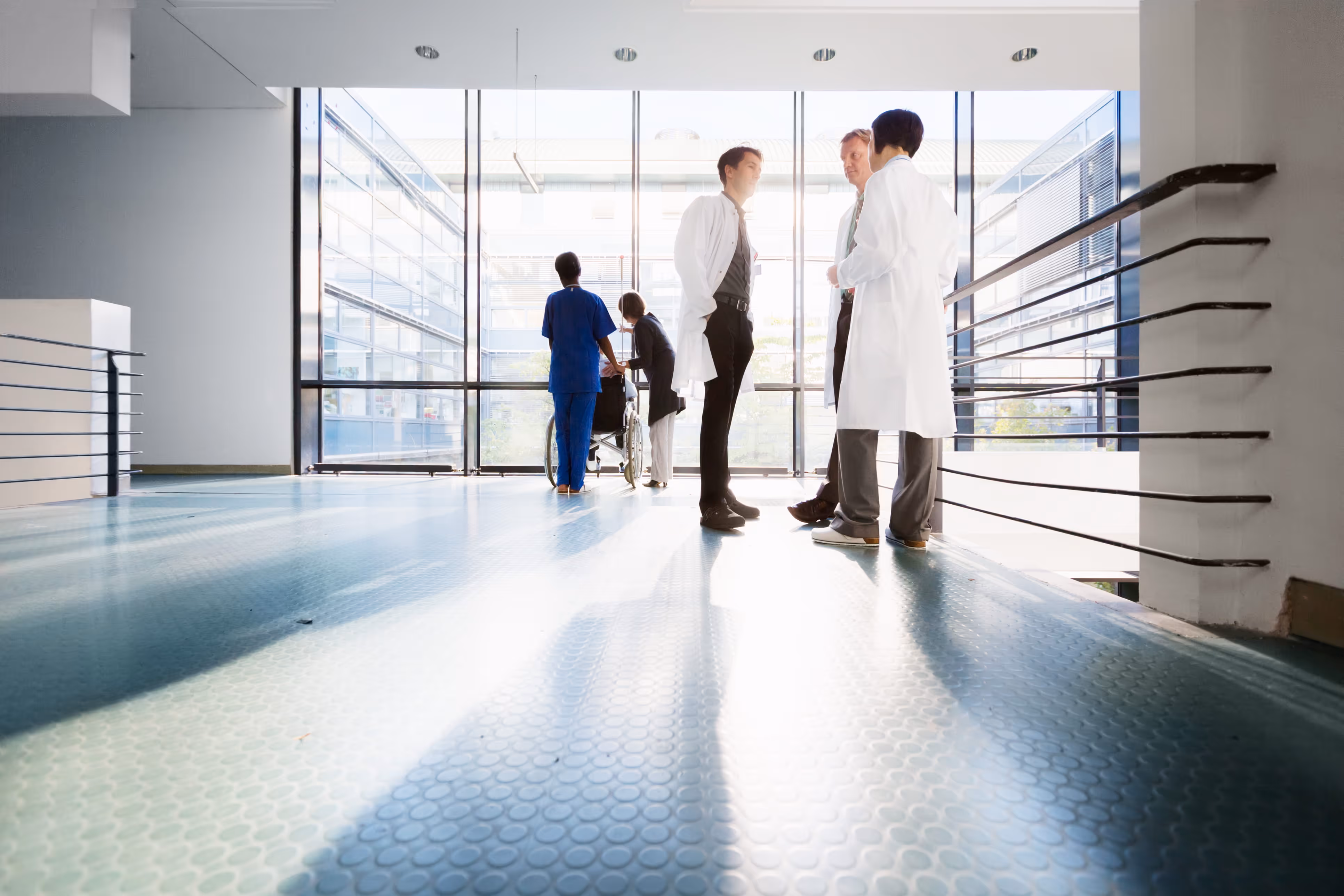 Medical staff in white coats and scrubs standing in a bright hospital hallway near large windows.