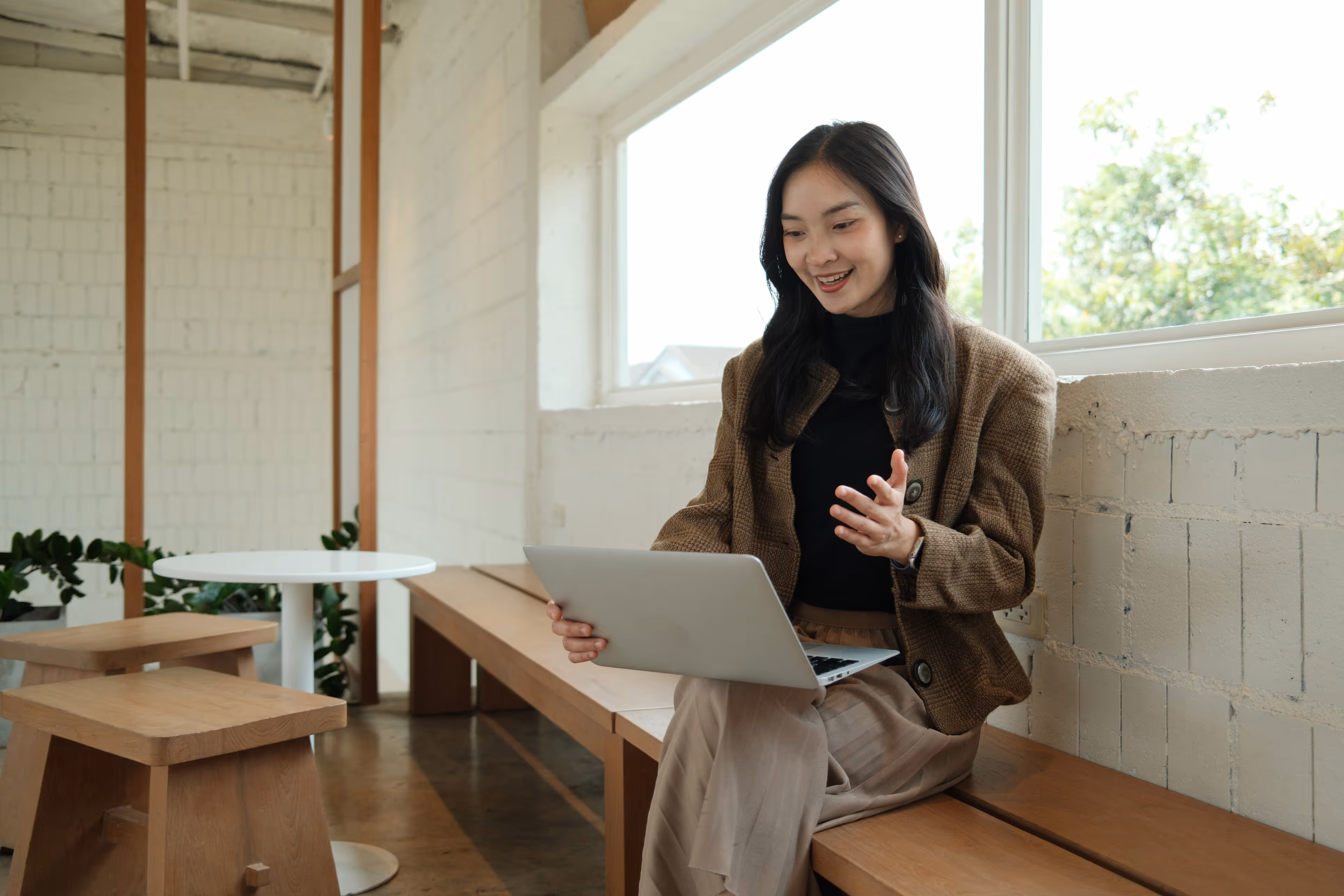Smiling woman sitting on a wooden bench using a laptop near a large window in a bright room.
