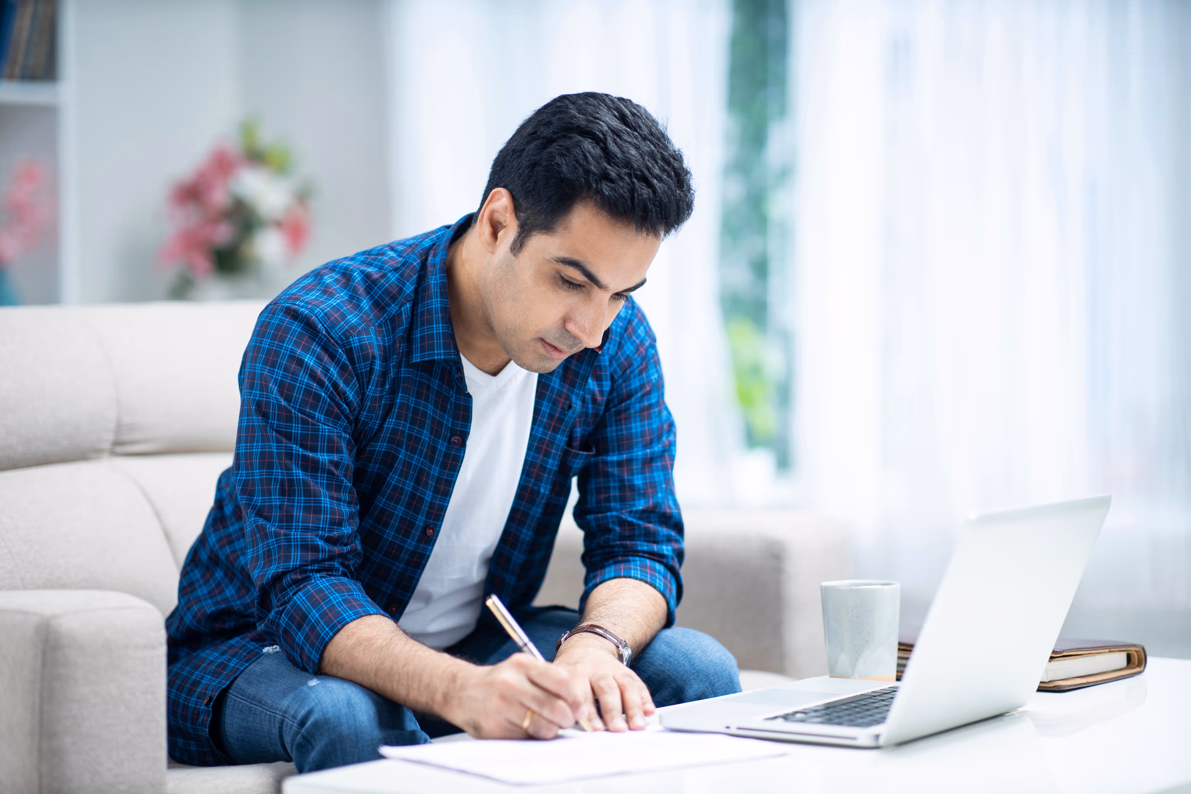 Man in blue plaid shirt writing on paper with an open laptop and mug on table nearby.