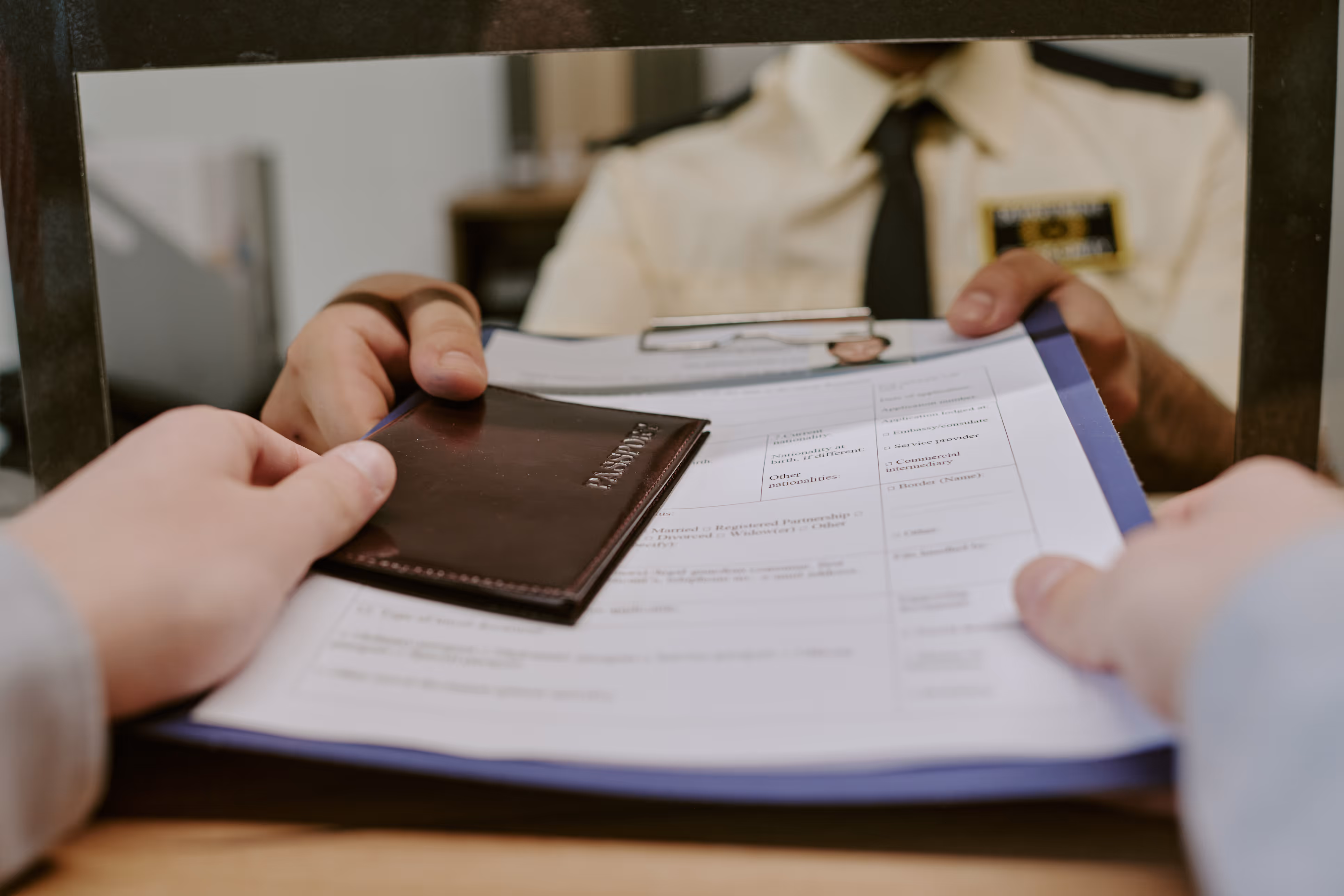 Person handing a passport and form to a uniformed officer through a service window.