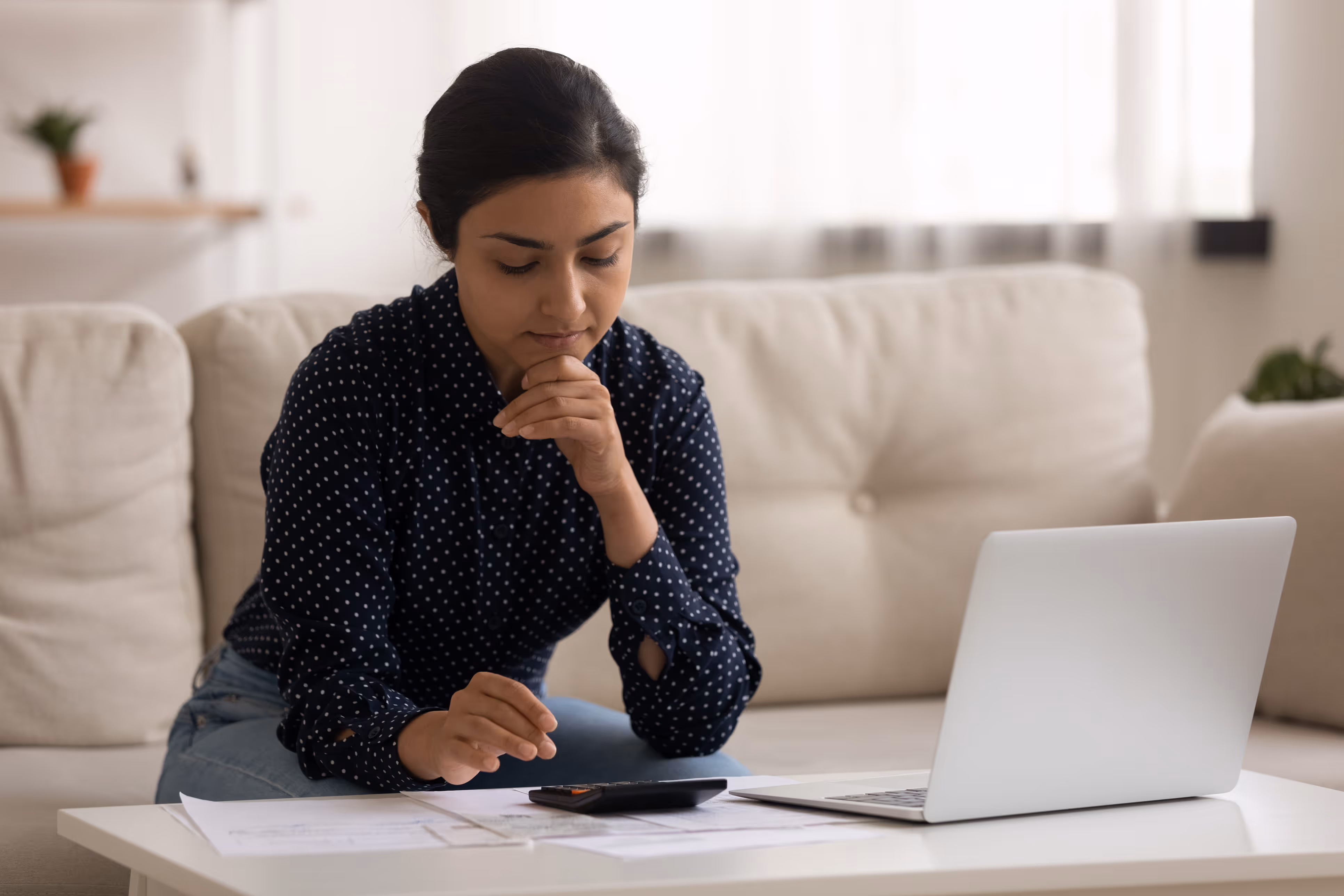 Woman sitting on a couch using a calculator with papers and a laptop on the table.