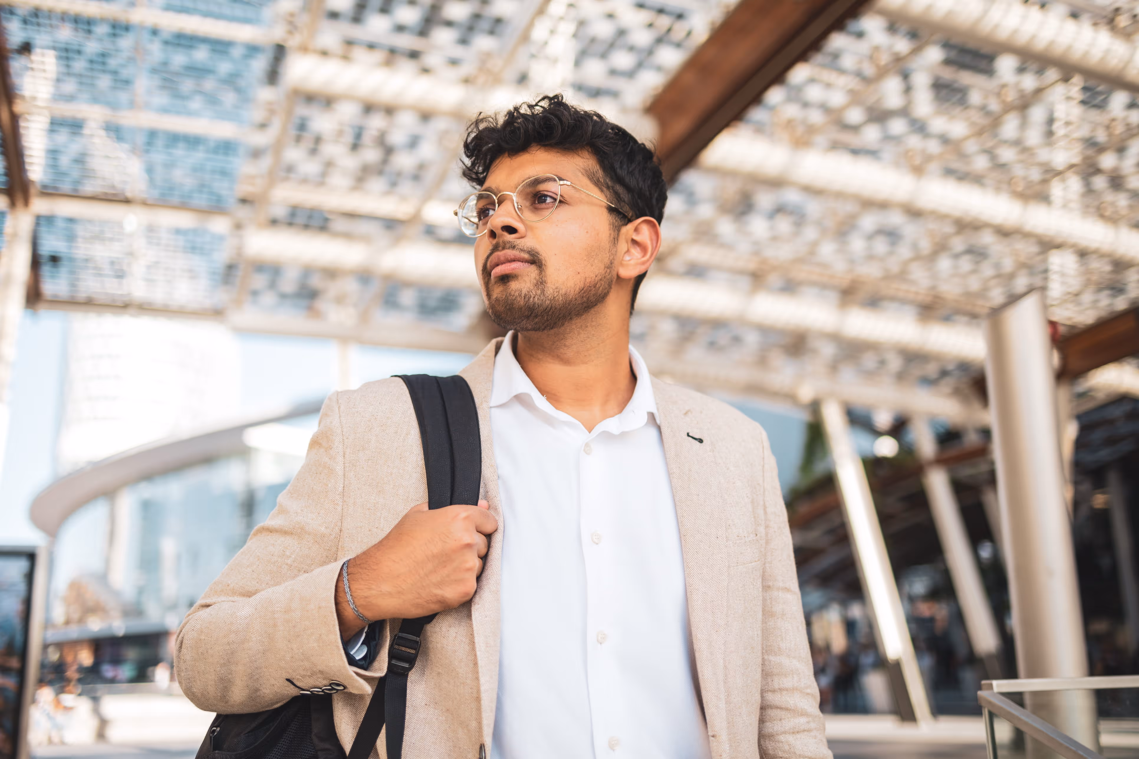 Confident man wearing glasses and beige blazer carrying backpack outdoors near modern architecture.