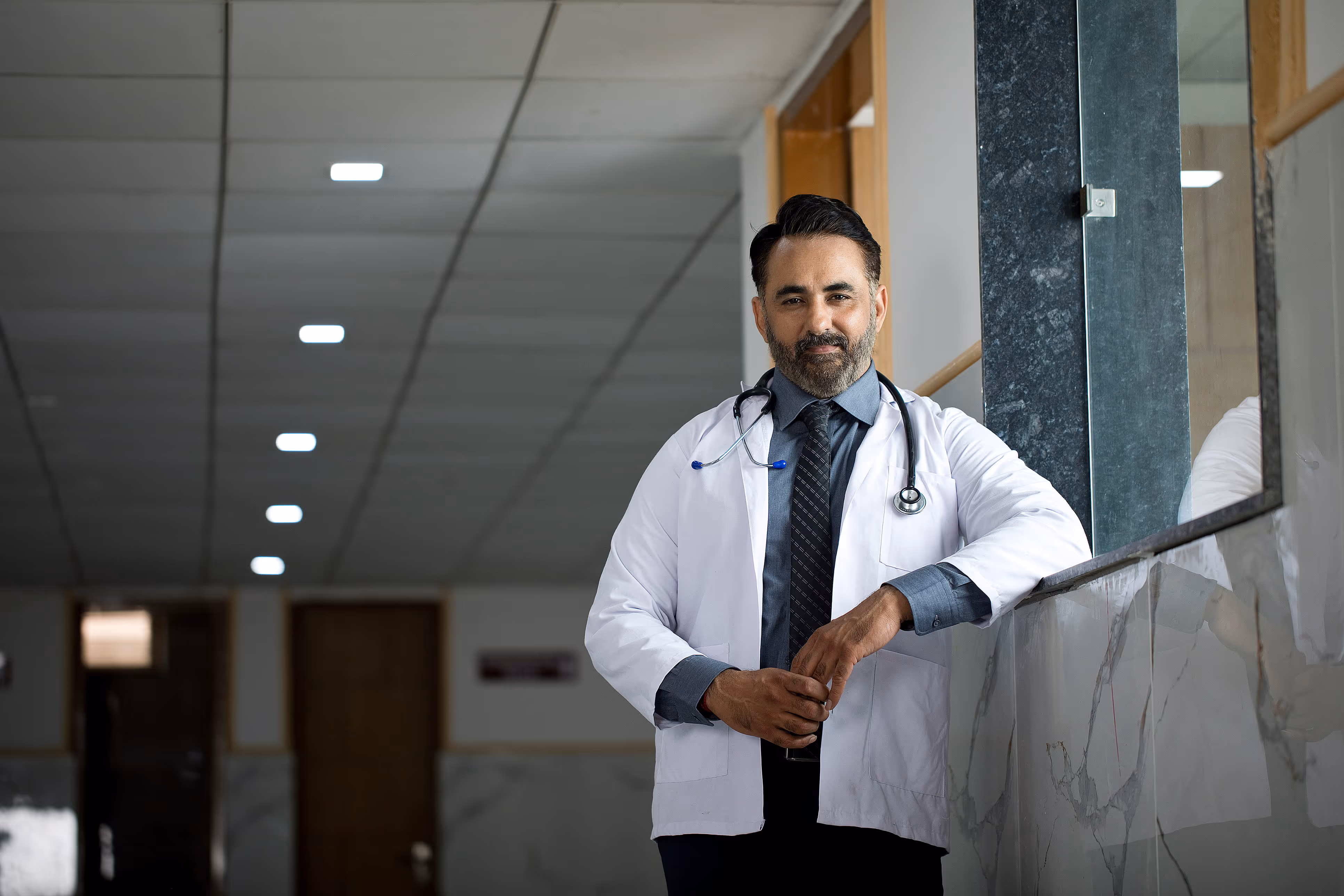 Confident male doctor in white coat with stethoscope leaning on marble ledge in hospital hallway.