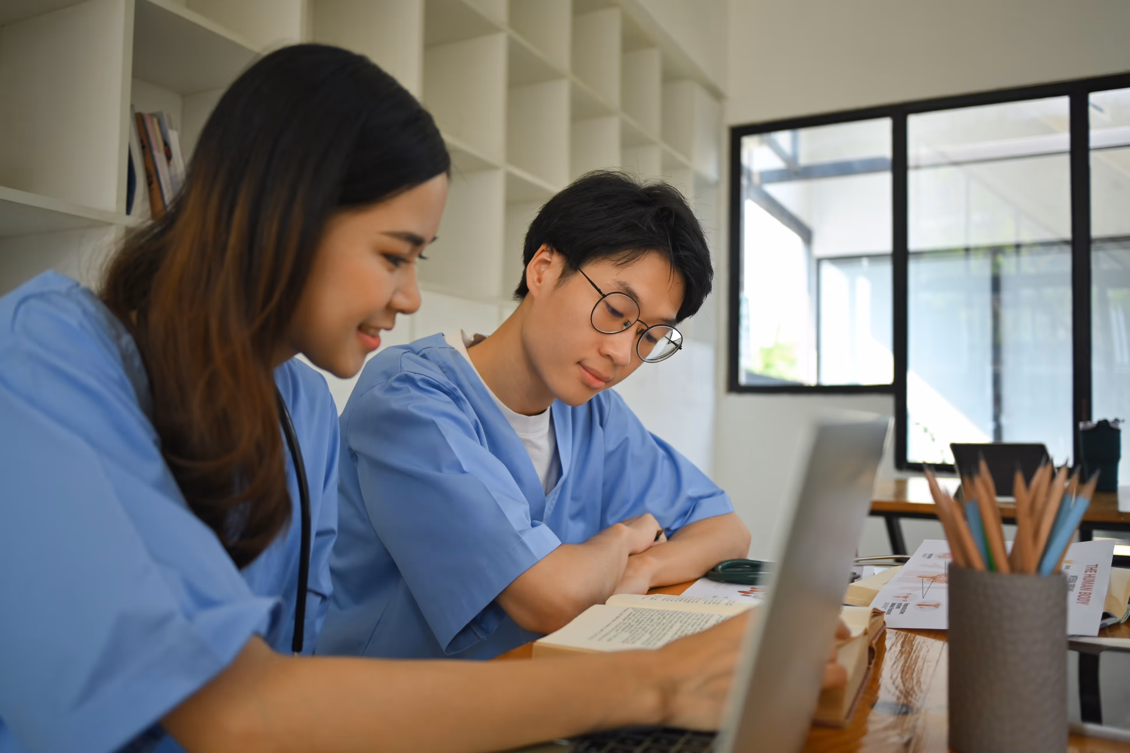 Two medical professionals in scrubs studying a book together at a table with a laptop nearby.