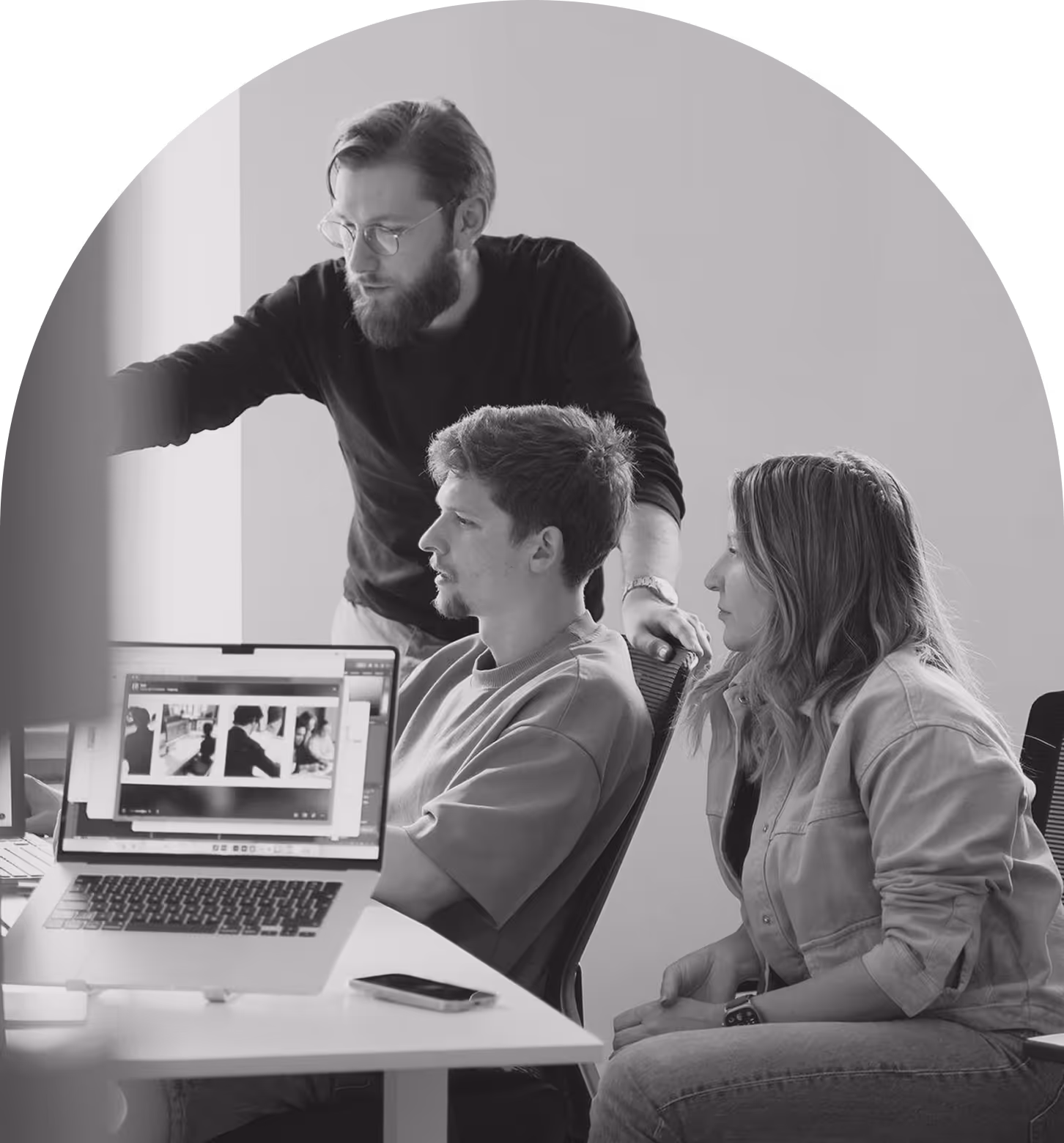 Three young people collaborating at a desk with a laptop showing photos on screen, one standing and pointing at the display.