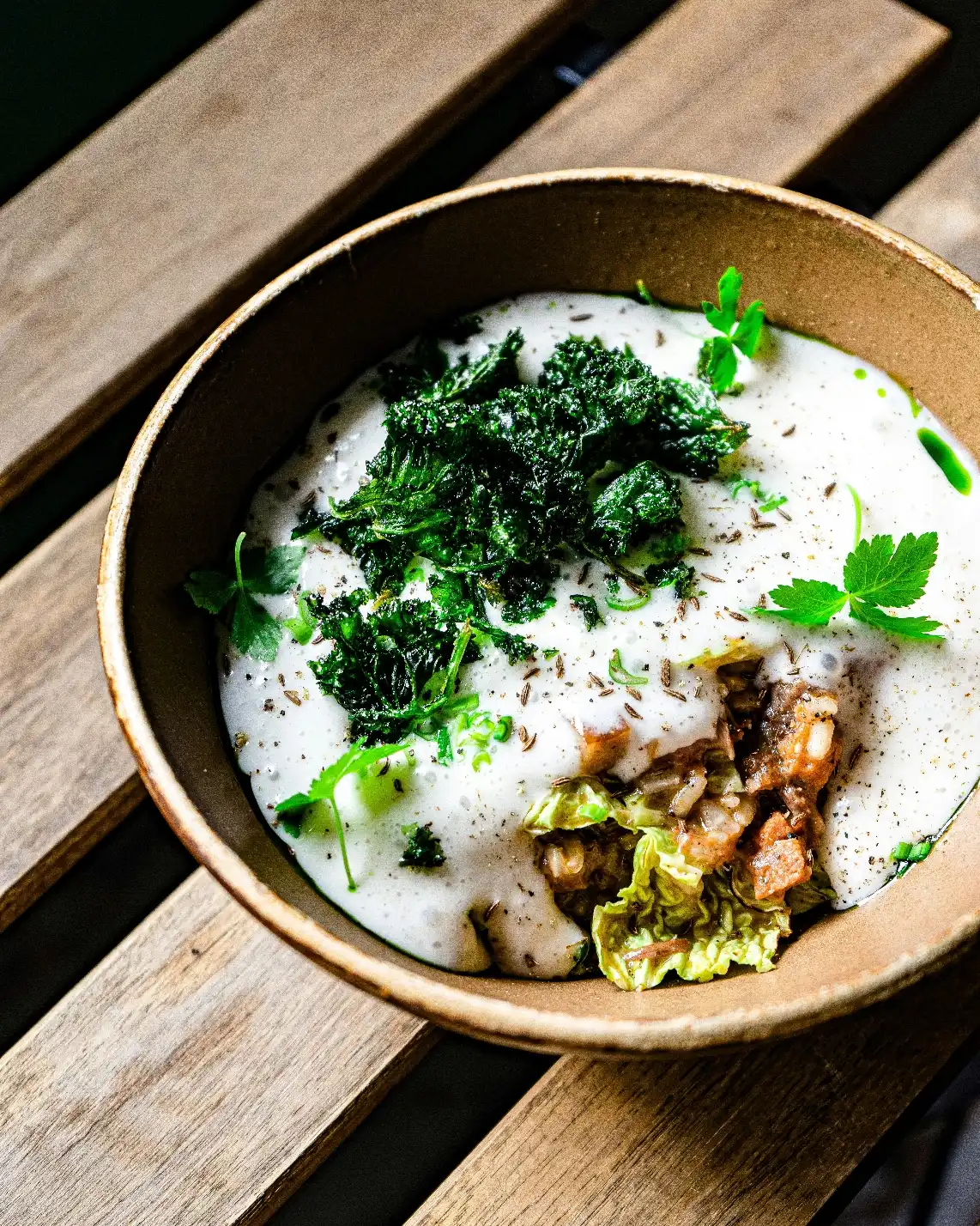 A creamy dish in a brown bowl with leafy greens and herbs, on a wooden table. The vibrant greens contrast with the rich, white sauce, creating a fresh, appetizing look.