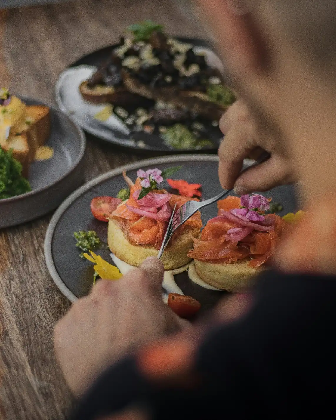 A person uses a fork and knife to eat a plate of gourmet brunch featuring smoked salmon on toast, garnished with flowers, with more dishes in the background.