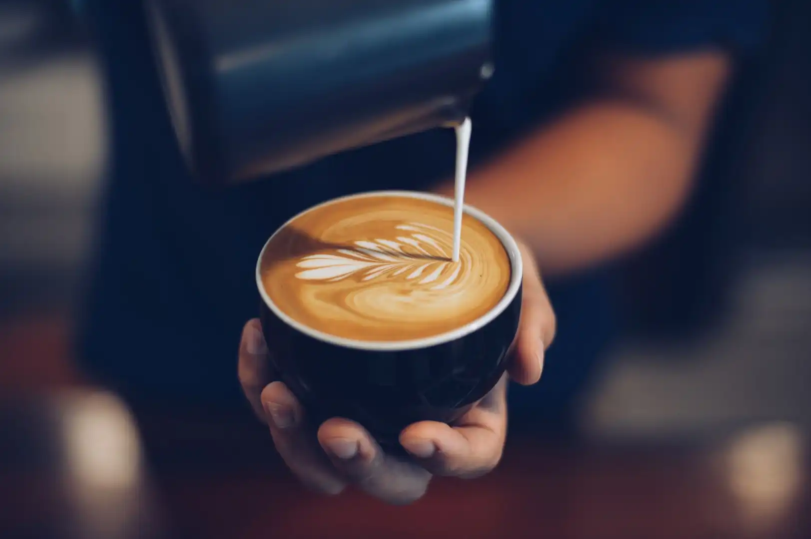 Close-up of a hand pouring milk to create artistic latte art with a leaf pattern in a black cup, conveying a cozy and skillful coffee-making atmosphere.