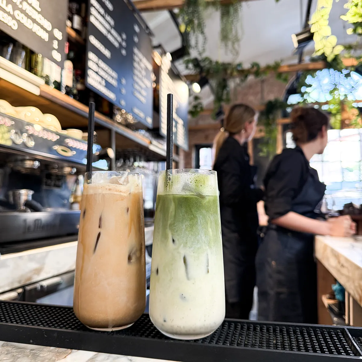 A row of cappuccinos in dark cups, each with heart-shaped latte art. Surrounded by potted green plants on a wooden table, creating a cosy café vibe.