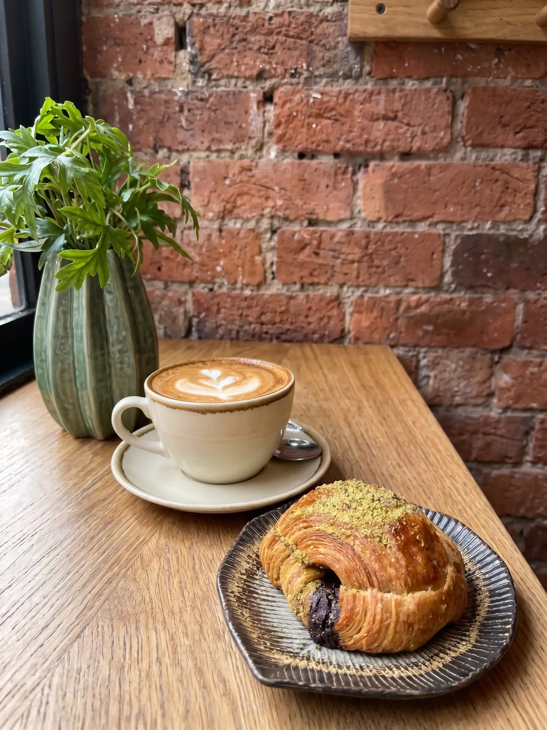 A rustic scone with berries sits on a dark plate next to a small black pot of cream. A silver spoon adds elegance, evoking a cozy and inviting atmosphere.