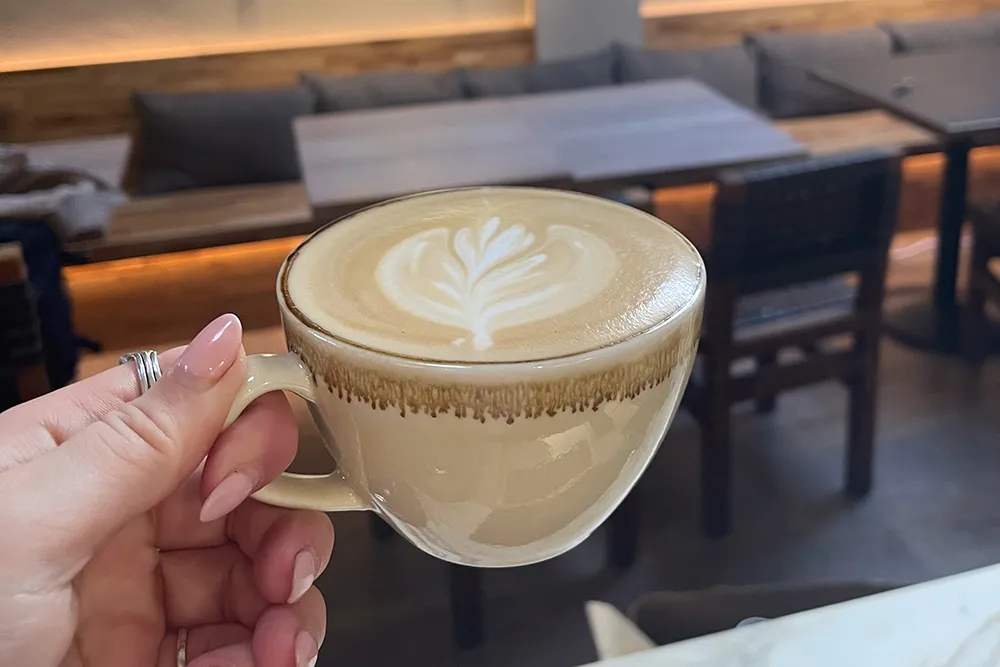 Close-up of a hand pouring milk to create artistic latte art with a leaf pattern in a black cup, conveying a cozy and skillful coffee-making atmosphere.