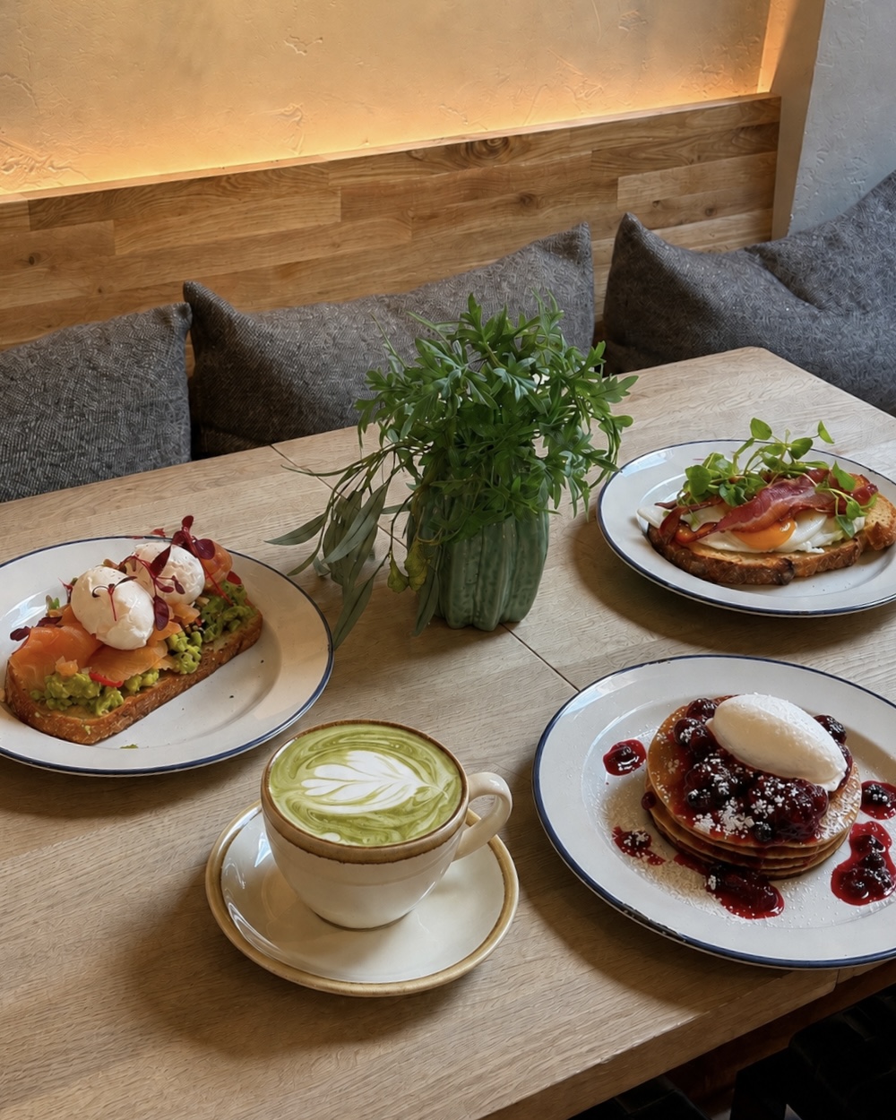 Cozy café table with matcha latte, three colorful brunch plates, leafy centerpiece, wood seating, and warm ambient lighting.