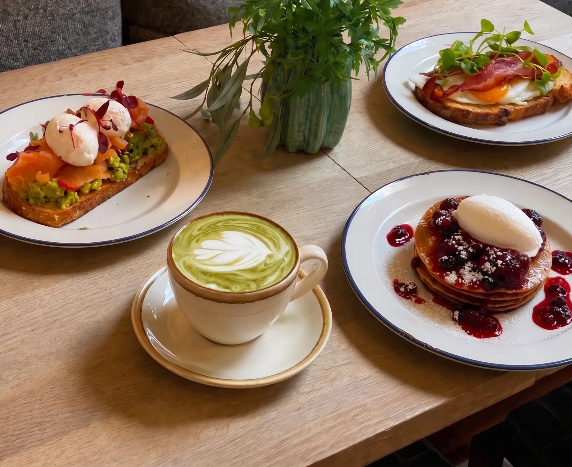 Cozy café table with matcha latte, three colorful brunch plates, leafy centerpiece, wood seating, and warm ambient lighting.