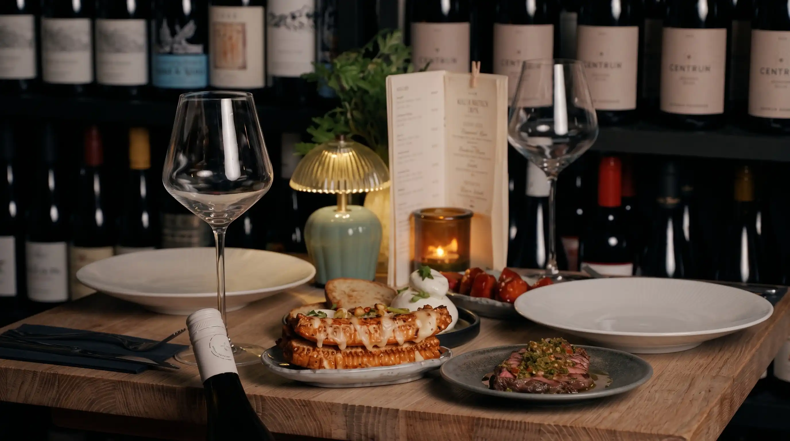 Restaurant table setting with two empty white plates, two empty wine glasses, a bottle of wine, a small lamp, a lit candle, a menu card, and plates of food including bread with melted cheese, burrata with garnish, grilled tomatoes, and sliced steak with green sauce, set against a background of wine bottles on shelves.