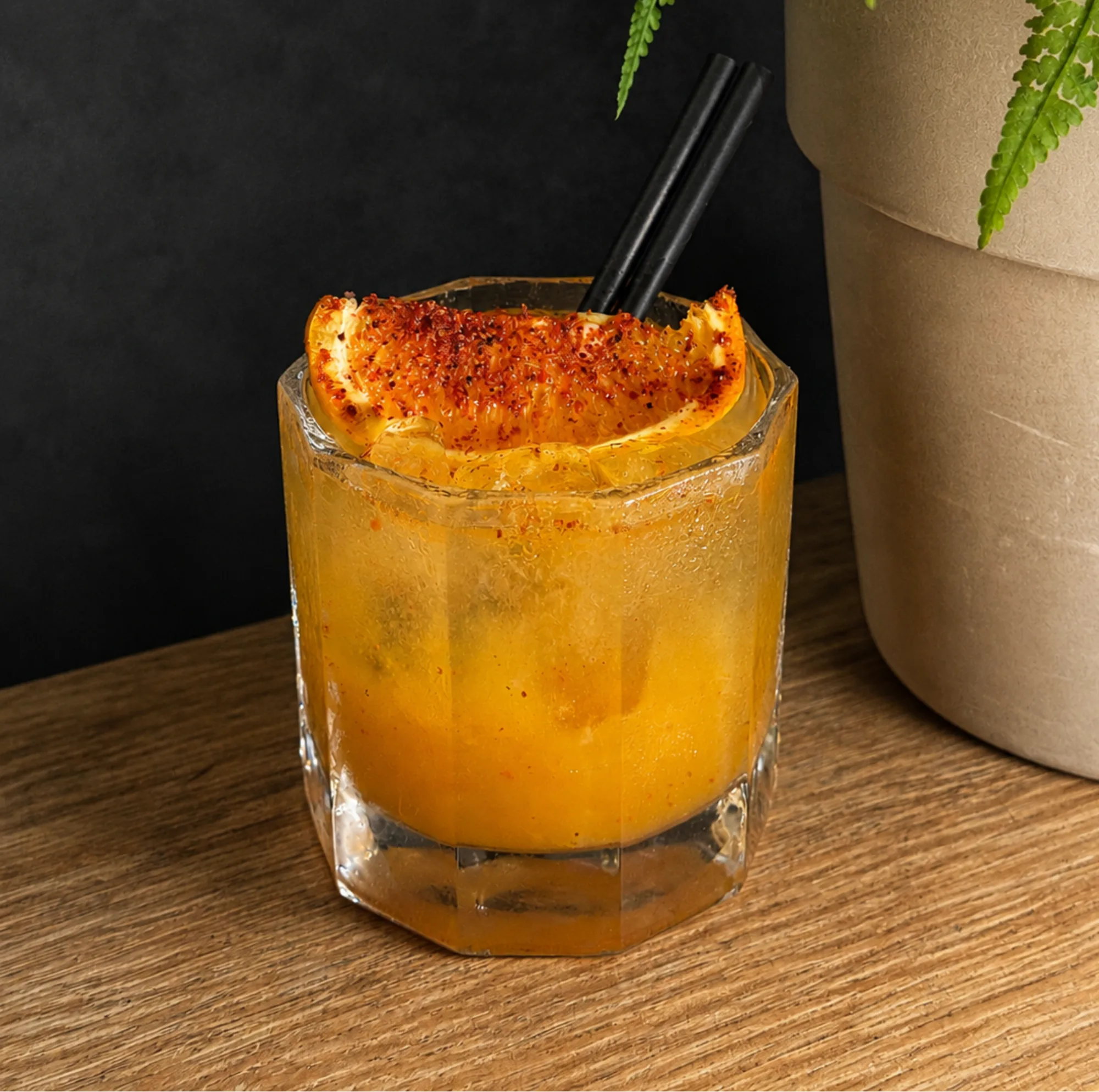 An orange cocktail over ice in a faceted glass, garnished with a chilli-dusted orange slice, beside a potted fern.