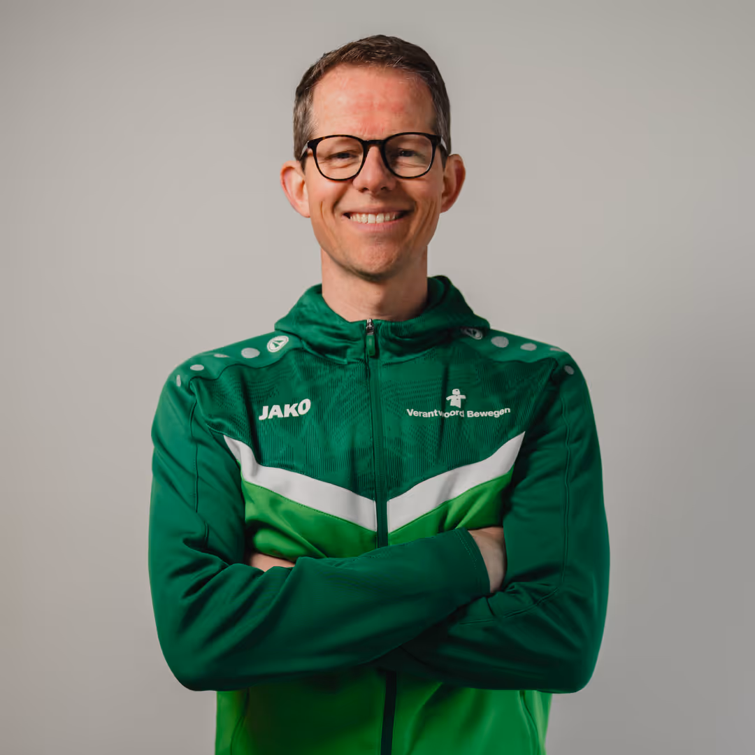 Smiling man with short hair and glasses wearing a green sports jacket with crossed arms against a plain background.