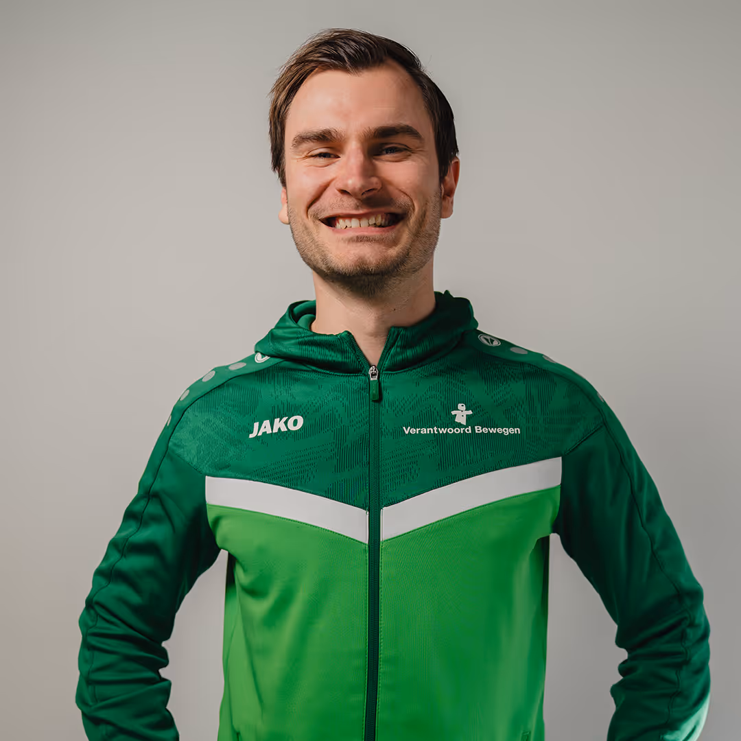 Smiling man wearing a green and dark green sports jacket with 'JAKO' and 'Verantwoord Bewegen' logos on a plain background.