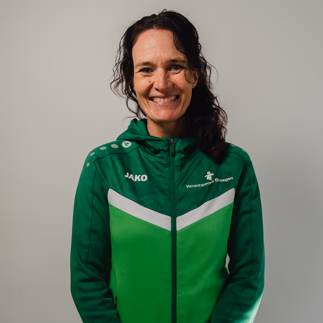 Woman with curly dark hair smiling, wearing a green zip-up sports jacket with 'JAKO' and 'Verantwoord Bewegen' logos.