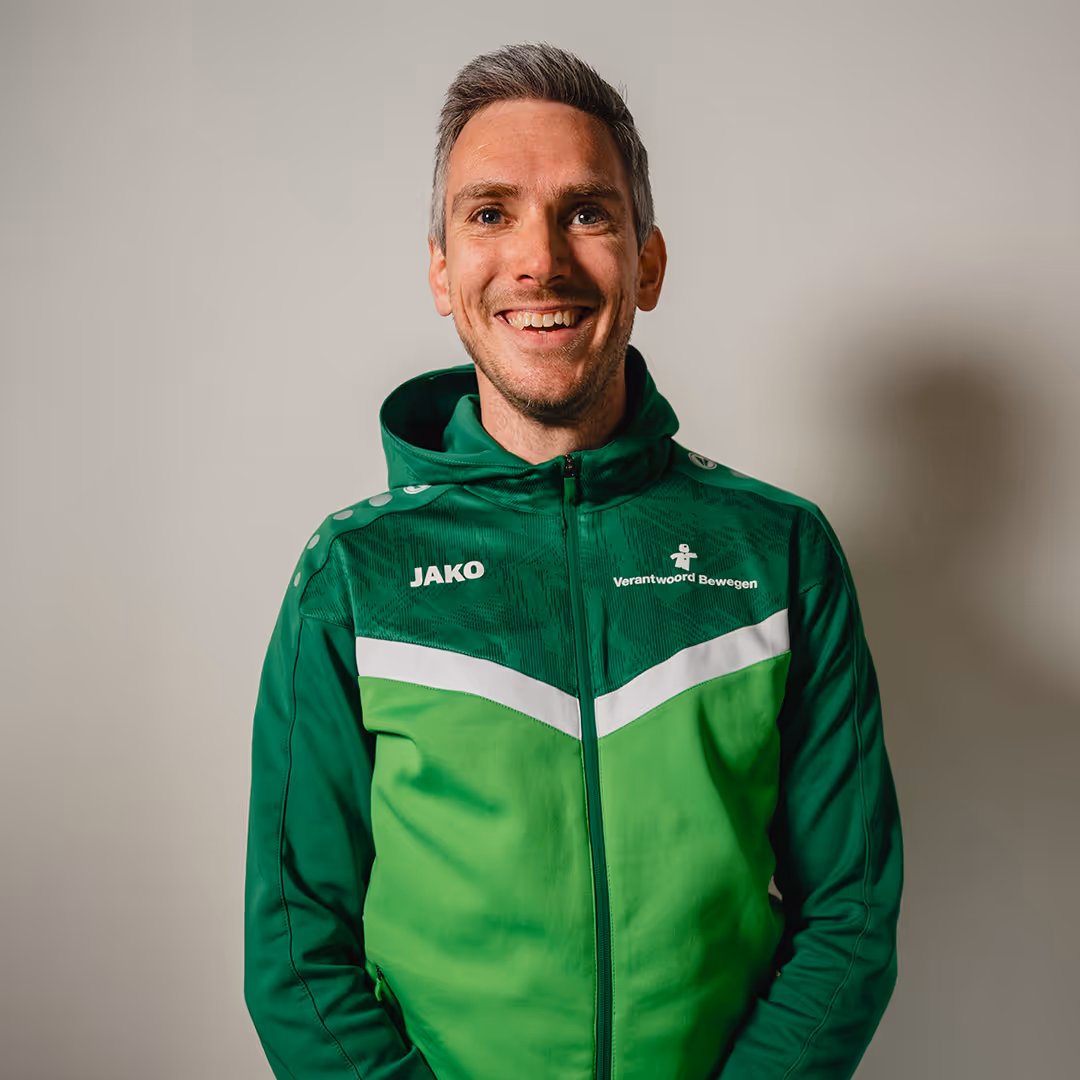Smiling man with short gray hair wearing a green JAKO sports jacket with 'Verantwoord Bewegen' logo on the chest.