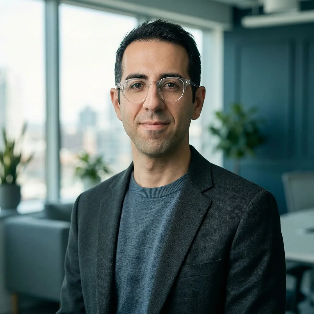 Man with glasses wearing a dark blazer and blue sweater in a modern office setting.