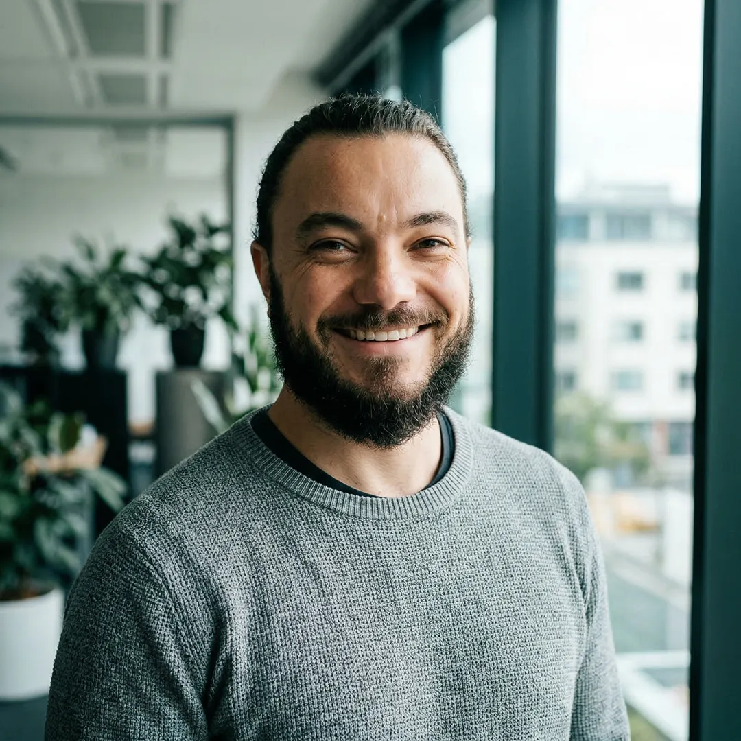 Smiling bearded man in gray sweater standing by a large window with plants and city view behind.