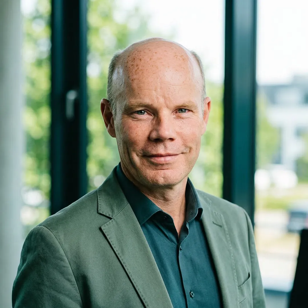 Portrait of a middle-aged bald man wearing a green jacket and dark shirt in a bright office.