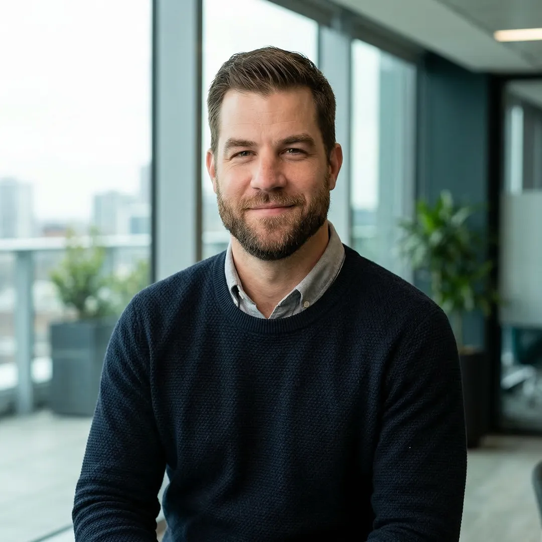 Smiling man with beard wearing a navy sweater and gray shirt in a modern office with large windows.