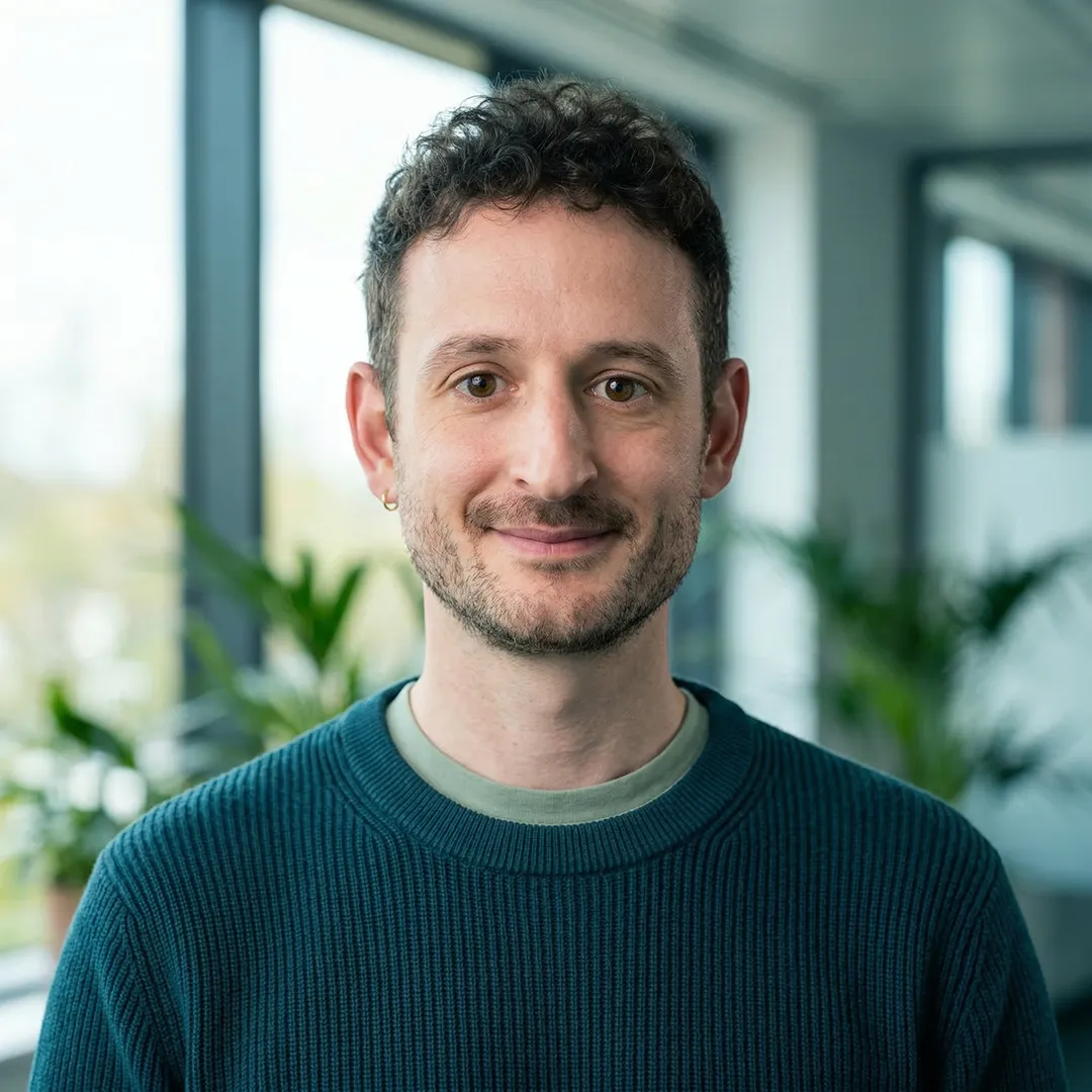 Smiling man with short curly hair and beard wearing teal sweater in modern indoor space with plants.