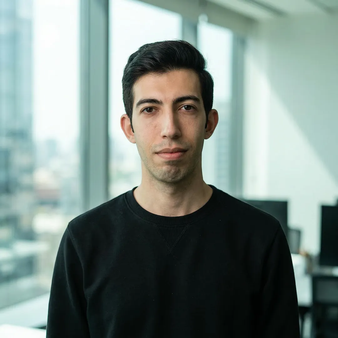 Portrait of a young man with black hair wearing a black sweater in a modern office with large windows.