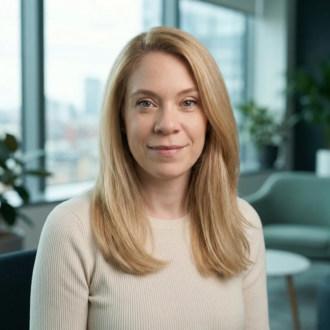 Smiling woman with light brown hair wearing a beige sweater in a modern office setting.