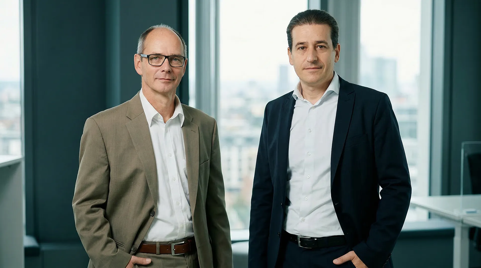 Two men in suits standing in an office with large windows overlooking a cityscape.