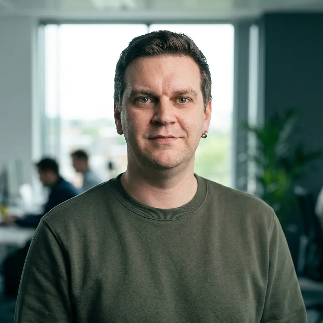 Man with short hair and earring wearing an olive green shirt in an office with blurred people behind.