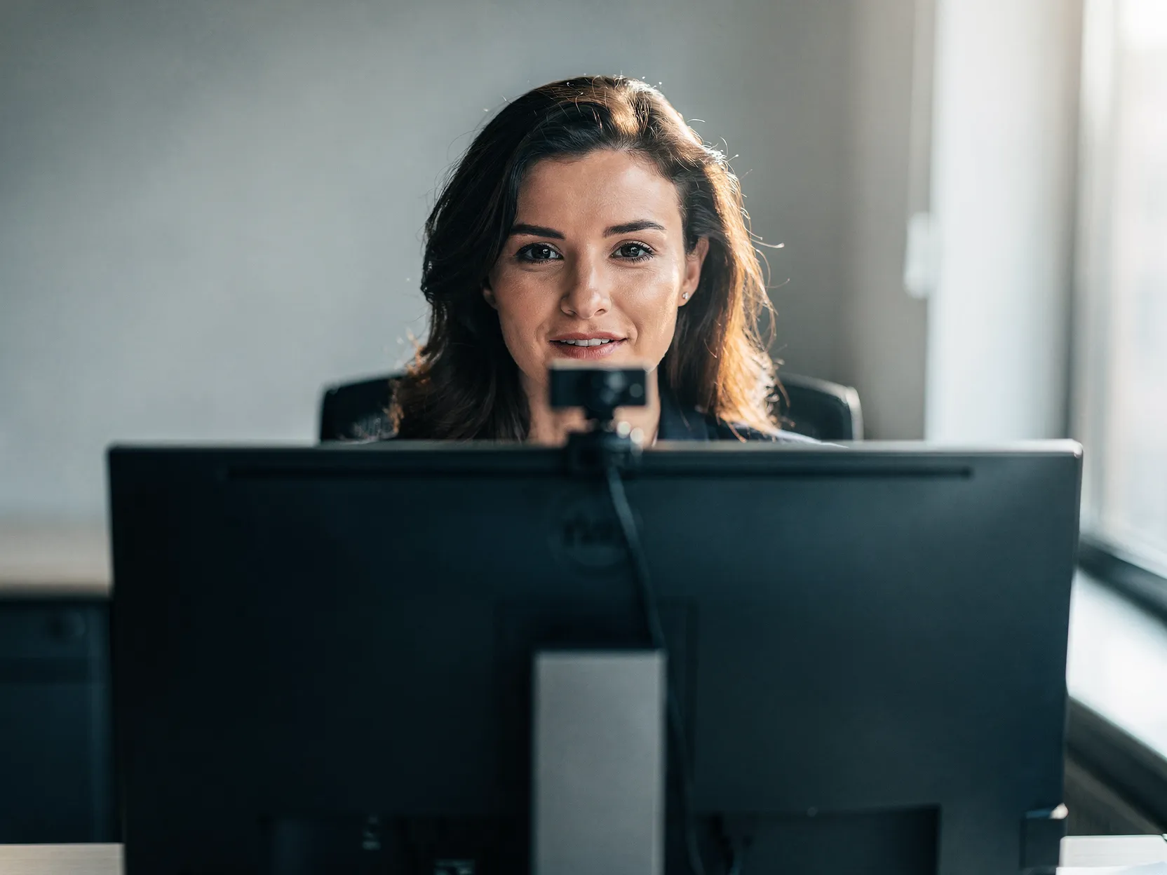 Woman sitting at a desk looking at a computer monitor with a webcam attached on top.